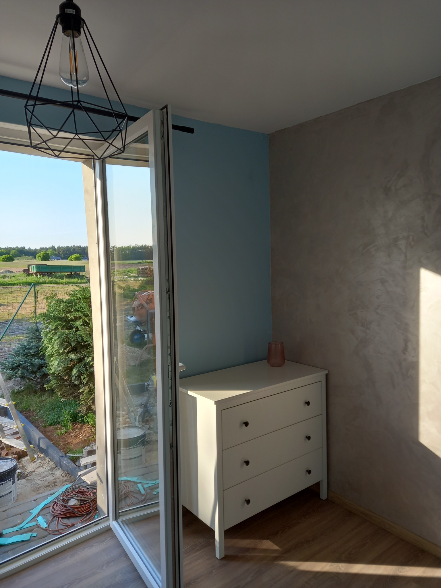 Wnętrze pokoju z otwartym oknem balkonowym, widoczna komoda z trzema szufladami, dekoracyjna ściana w odcieniach szarości i błękitu, na zewnątrz widok na ogród i teren budowy.