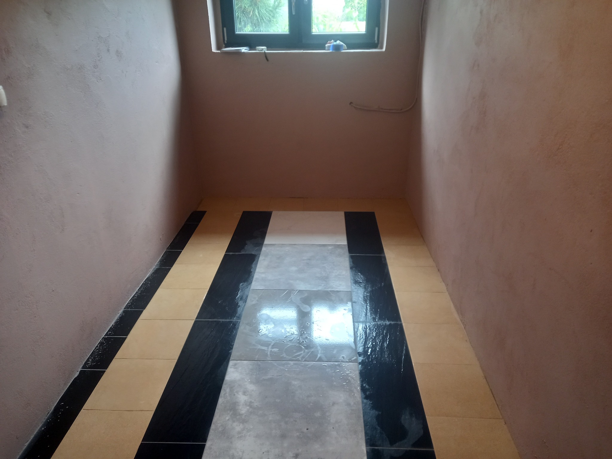 Wnętrze łazienki z ułożonymi płytkami podłogowymi w odcieniach beżu, szarości i czerni, ściany w kolorze brudnego różu, małe okno z widokiem na zewnątrz.