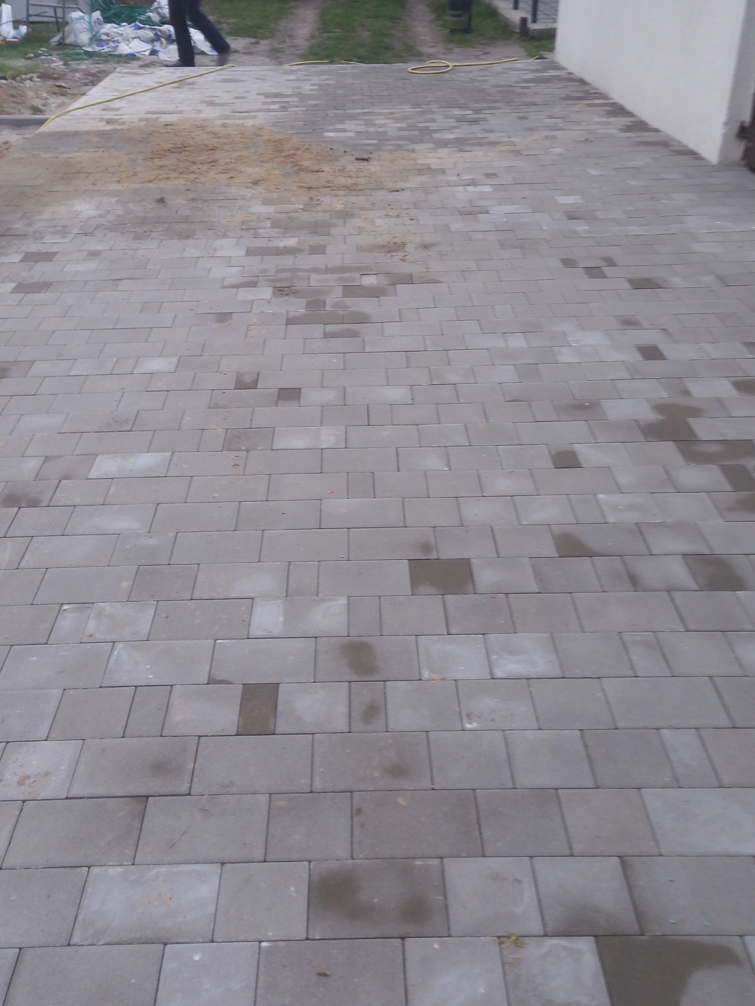 Ułożona szara kostka brukowa na podjeździe, widoczne ślady wilgoci i piasku, w tle częściowo widoczny budynek.