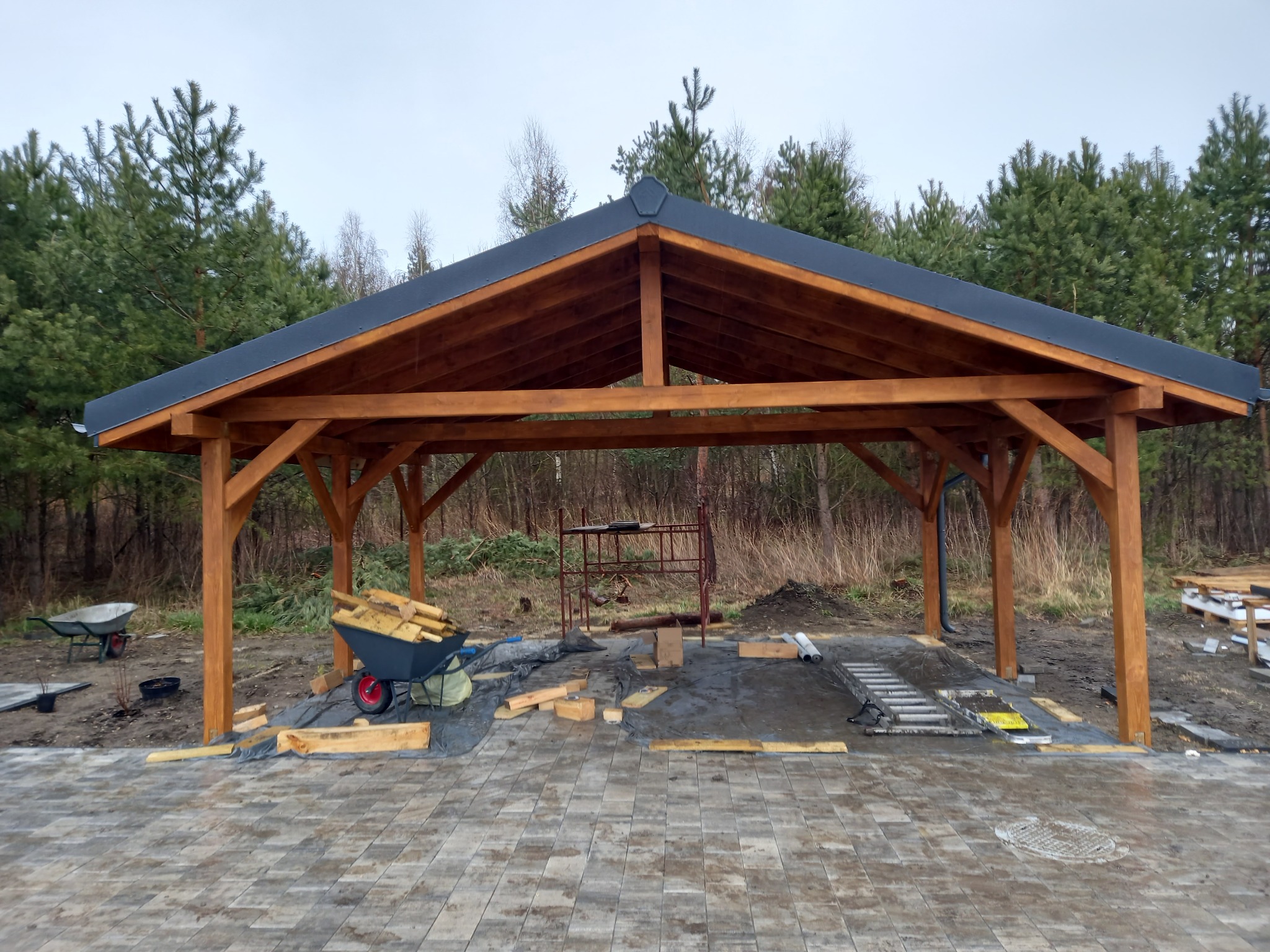 Drewniana konstrukcja z czarnym dachem na tle lasu, wózek z drewnem i rozłożone narzędzia na kostce brukowej, częściowo ukończona budowa.