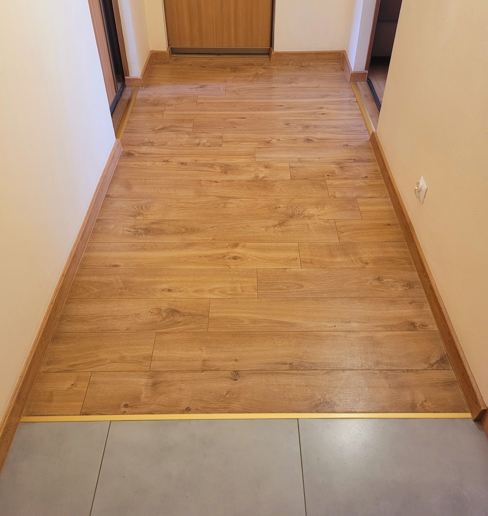 Ułożone panele podłogowe w korytarzu, widoczne przejście z płytek, listwy przypodłogowe w kolorze paneli, drzwi w tle.