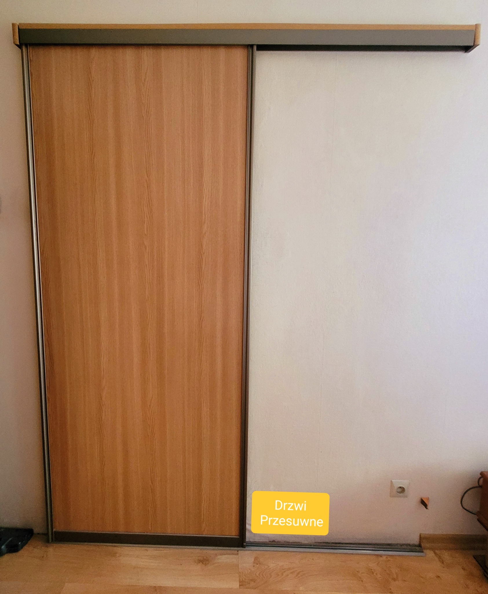 Częściowo przesunięte drzwi przesuwne szafy wnękowej z drewnopodobnym panelem i białym wypełnieniem, widoczna szyna i fragment podłogi.