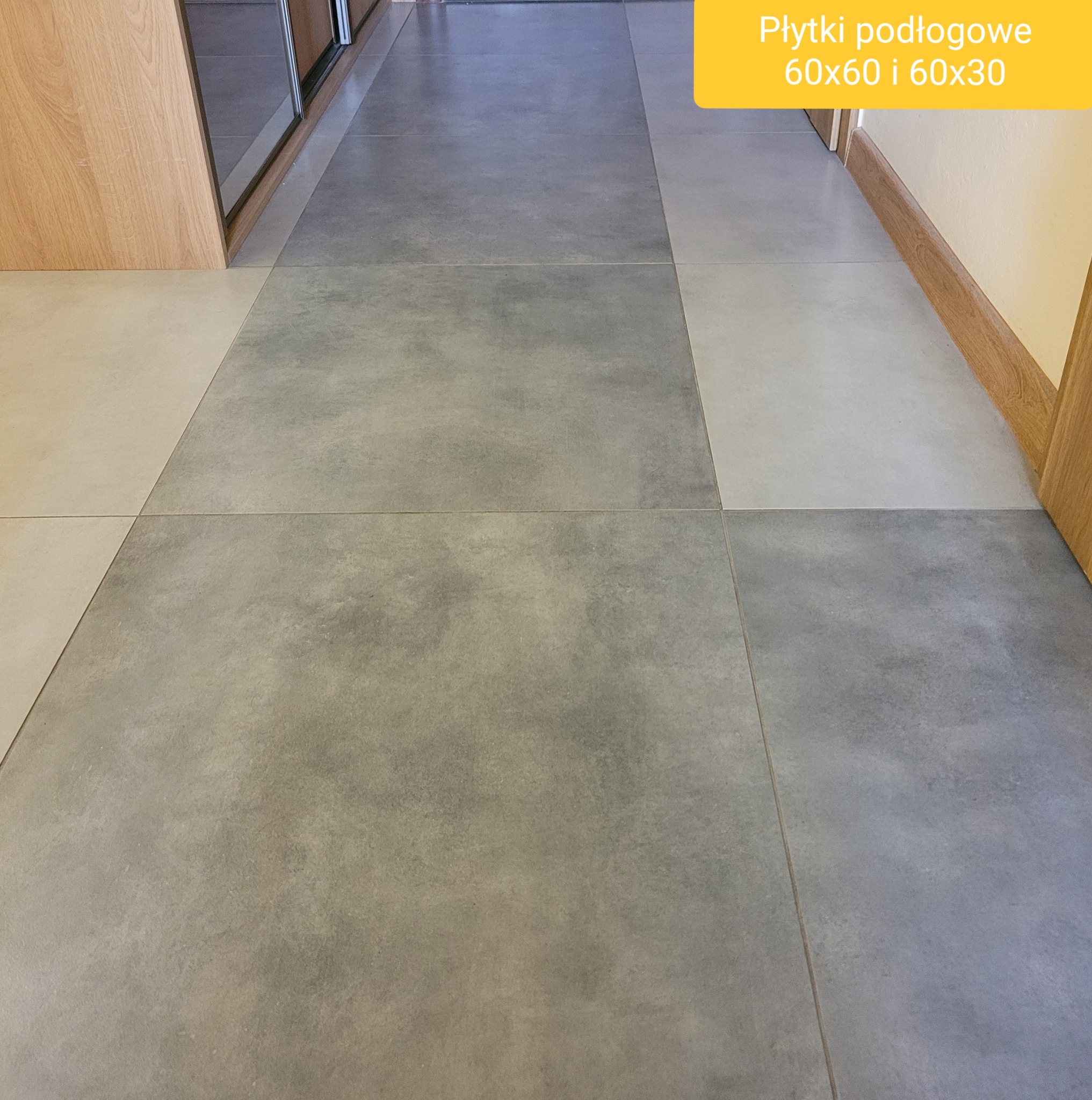 Szare i beżowe płytki podłogowe ułożone w korytarzu, widoczne drewniane listwy przypodłogowe i fragment szafy w zabudowie.