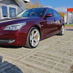 2brodetailing - Czerwony, lśniący samochód BMW serii 5 po umyciu, zaparkowany na placu z betonowej kostki, z widoczną myjnią samochodową w tle.