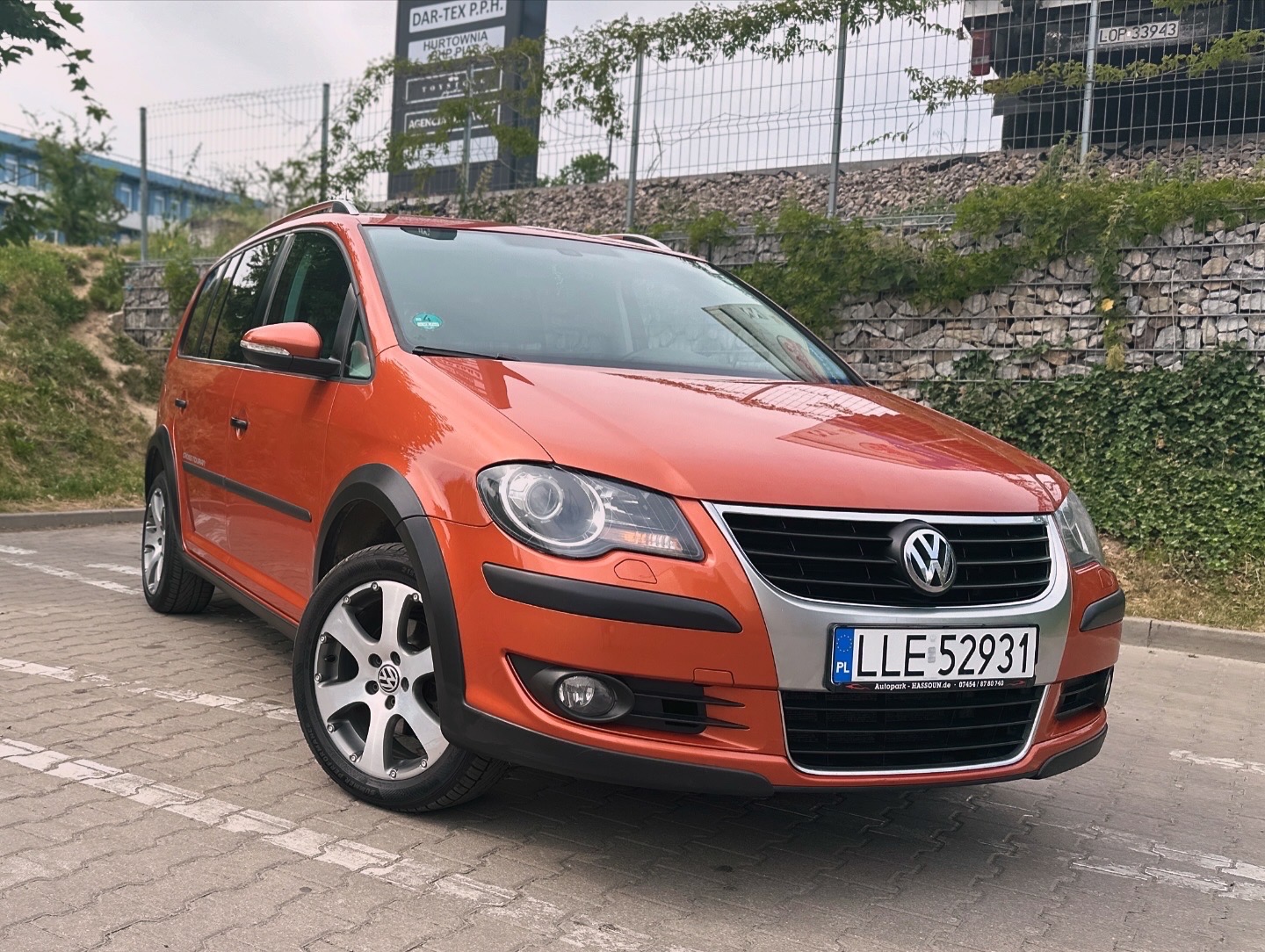 Pomarańczowy Volkswagen Touran Cross na kostce brukowej, widok z przodu, z widoczną tablicą rejestracyjną i logiem VW na feldze.