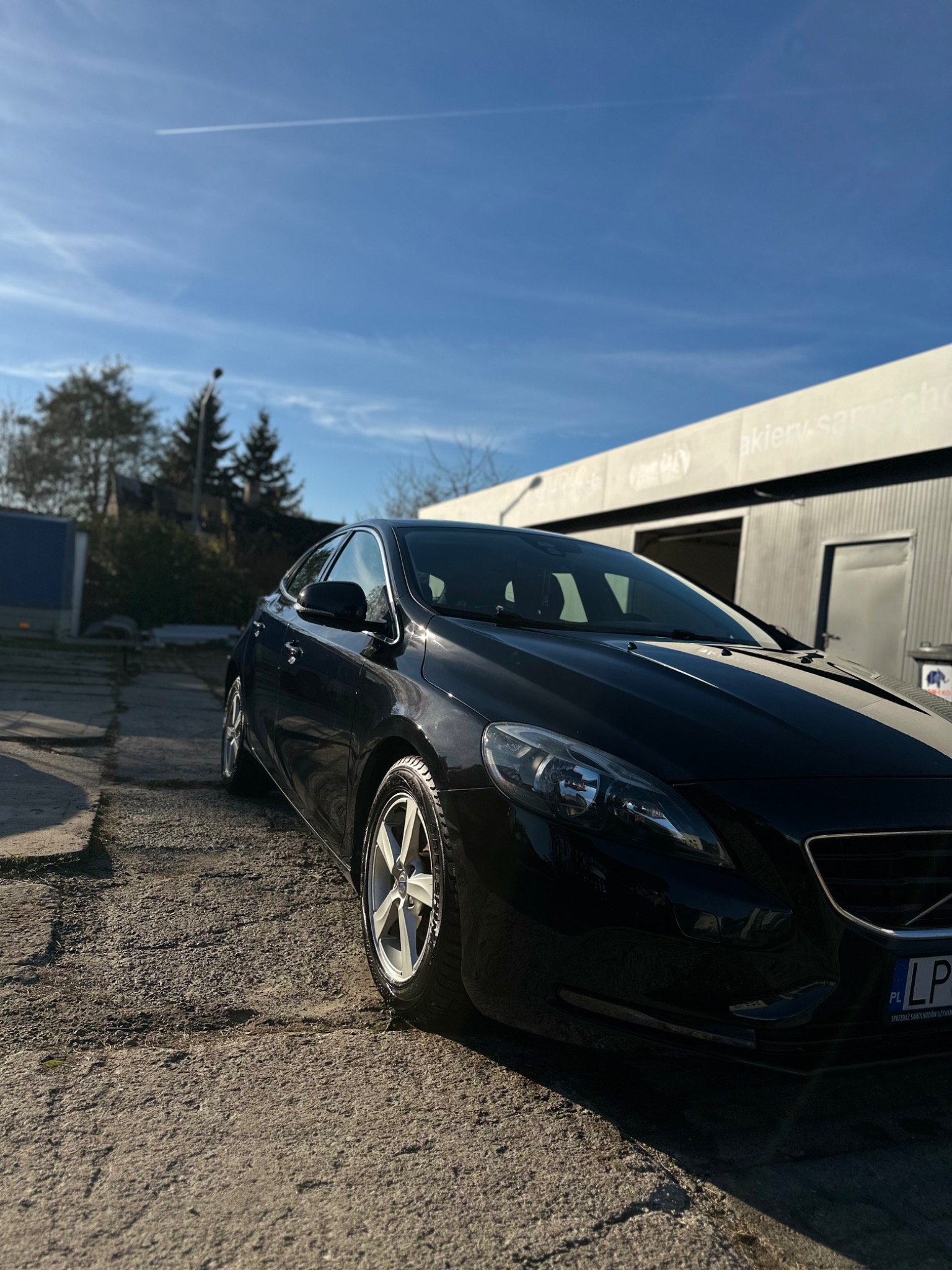 Czarny samochód osobowy marki Volvo, zaparkowany przed budynkiem myjni w słoneczny dzień, widoczny numer rejestracyjny.