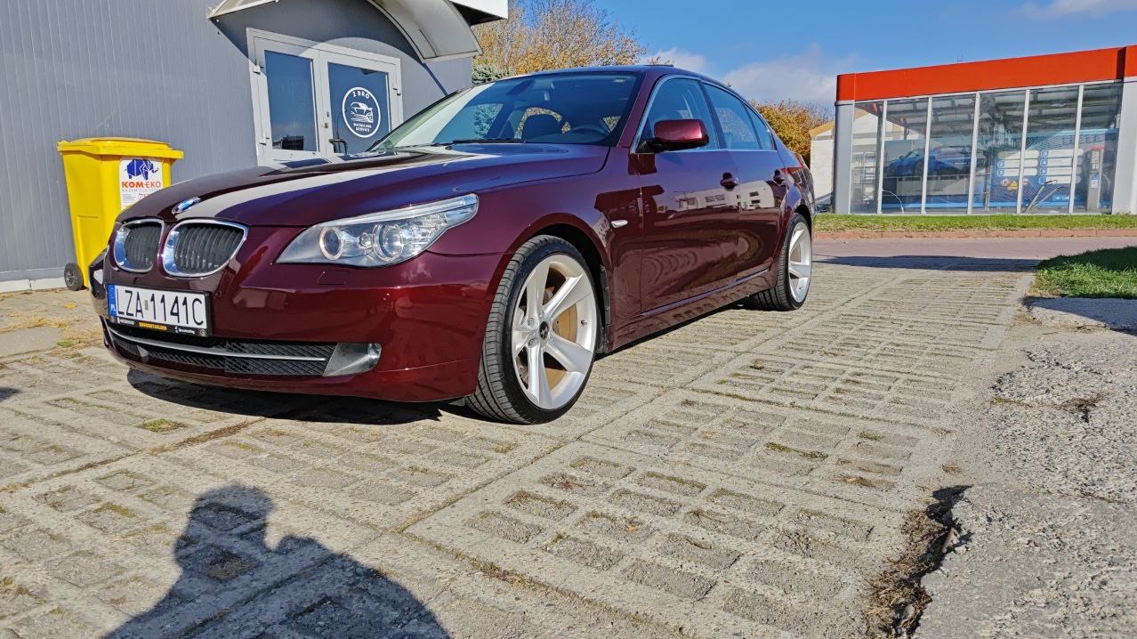 Czerwony, lśniący samochód BMW serii 5 po umyciu, zaparkowany na placu z betonowej kostki, z widoczną myjnią samochodową w tle.