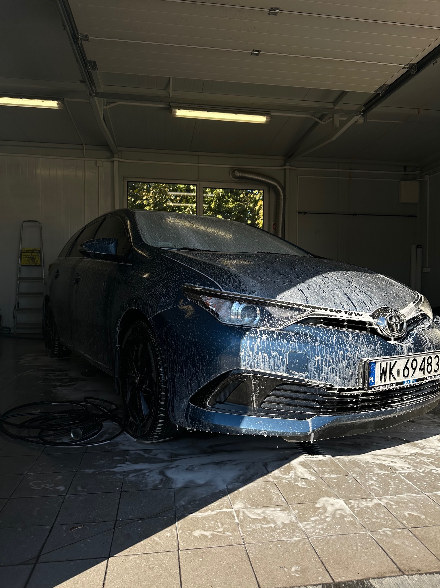 Niebieski samochód pokryty pianą w myjni, widoczne czarne felgi i przewód od myjki ciśnieniowej leżący na posadzce.