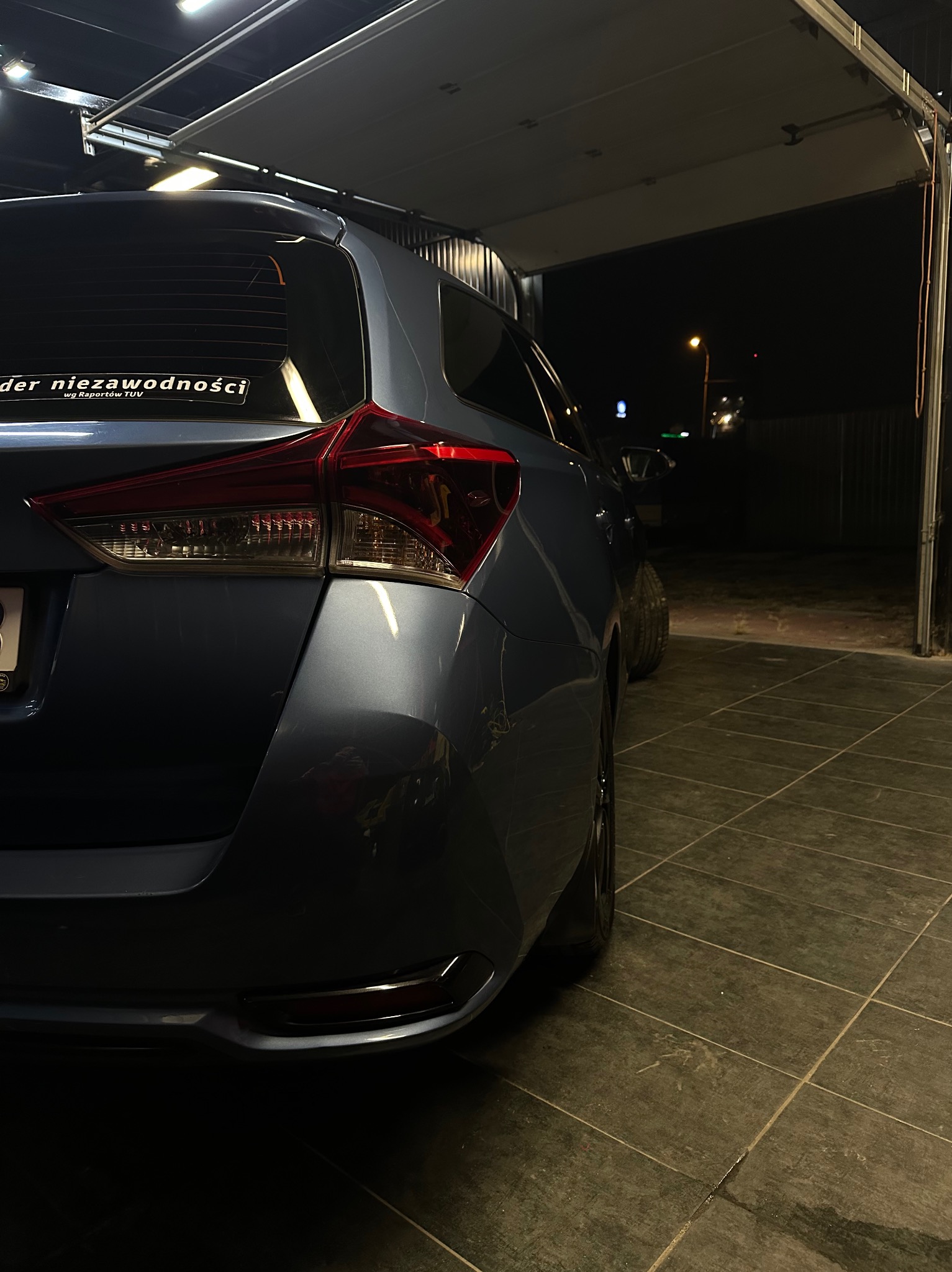 Błękitny samochód typu kombi, widok z tyłu, zaparkowany na kafelkowanej posadzce w garażu z otwartymi drzwiami, naklejka 'lider niezawodności' na tylnej szybie.