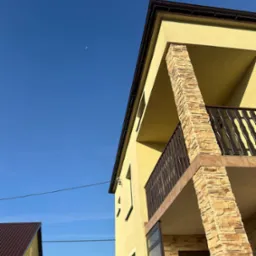 Żółta elewacja domu z balkonem wykończonym kamieniem dekoracyjnym w słoneczny dzień, widok z dołu na tle błękitnego nieba z widocznym księżycem.