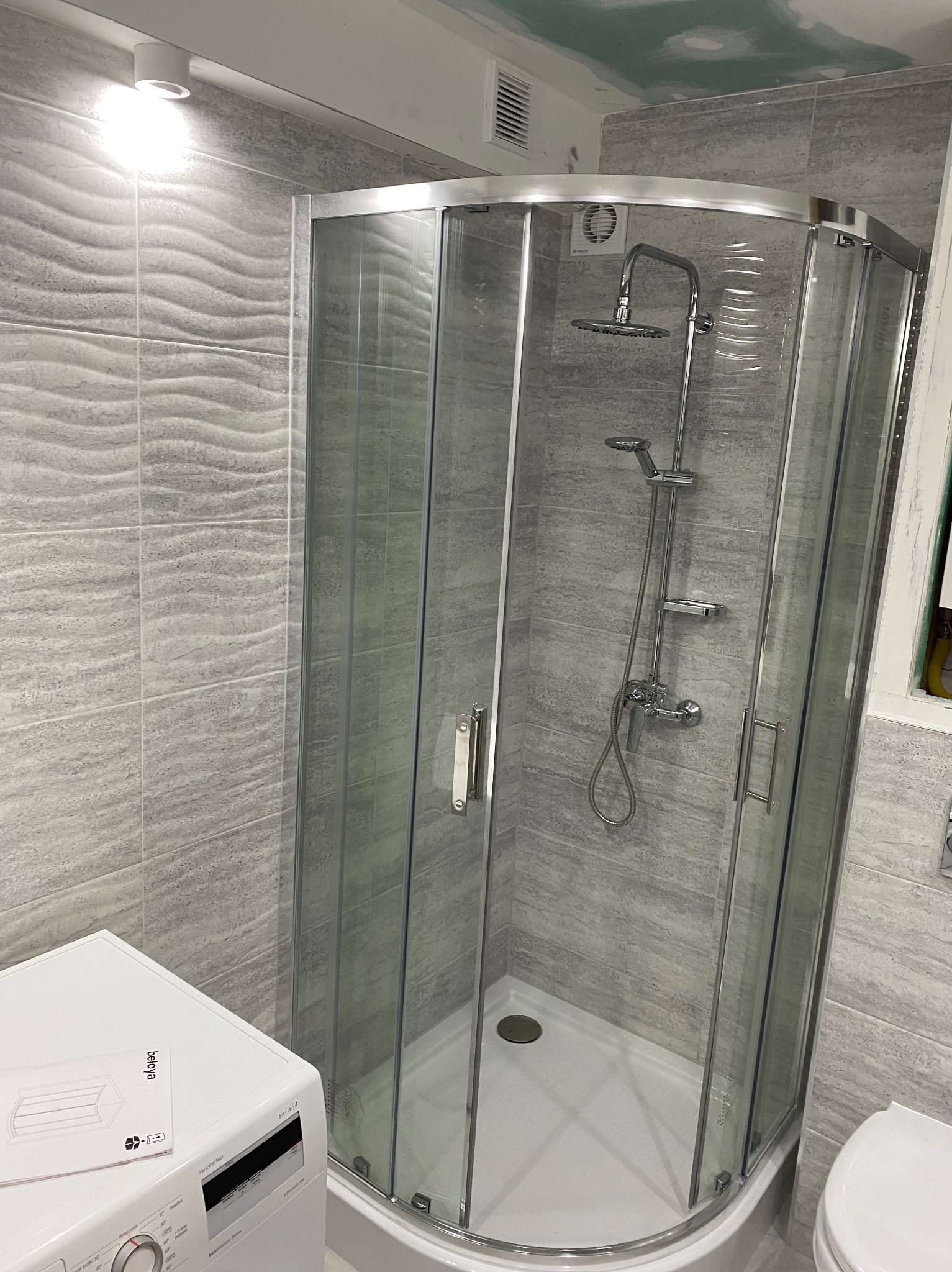 Szklana kabina prysznicowa narożna z chromowanymi profilami i deszczownicą zamontowana w łazience wykończonej szarymi płytkami imitującymi kamień, obok widoczna pralka z instrukcją obsługi.