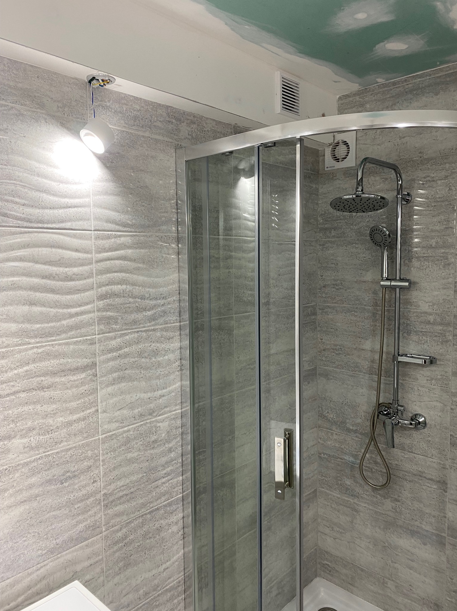 Szara łazienka z kabiną prysznicową z deszczownicą, oświetleniem punktowym i wentylacją, wykończona płytkami imitującymi kamień z falistą strukturą, sufit częściowo wykończony płytami...