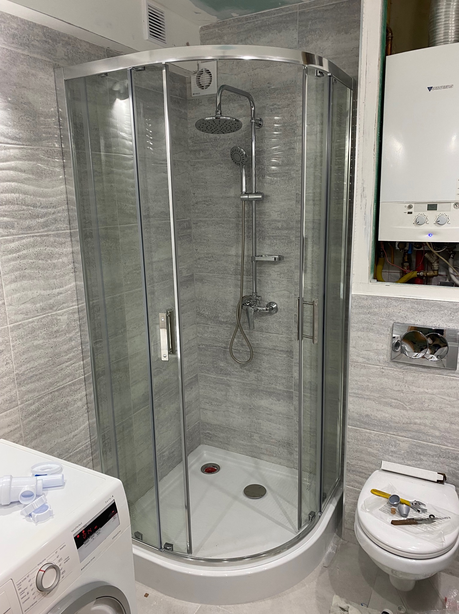 Nowoczesna kabina prysznicowa z chromowanymi profilami i deszczownicą, szare płytki imitujące kamień na ścianach, pralka i toaleta w tle, widoczne narzędzia i elementy instalacyjne.