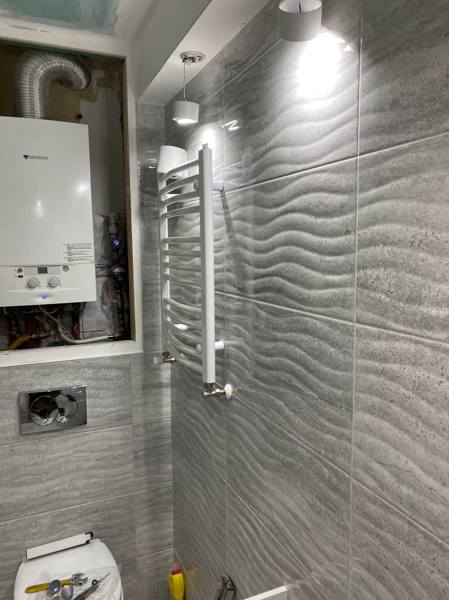 Szare płytki imitujące falę na ścianie w łazience, biały grzejnik drabinkowy, wiszący piec gazowy w zabudowie, narzędzia na sedesie.