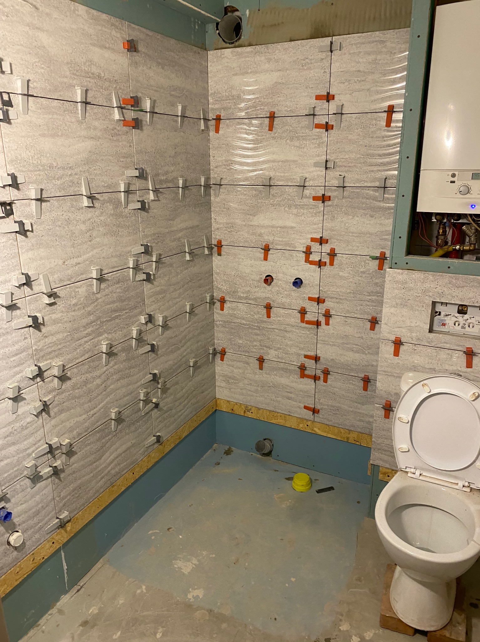 Łazienka w trakcie kładzenia płytek ściennych, z użyciem systemów poziomowania, widoczny bojler i toaleta w pomieszczeniu.