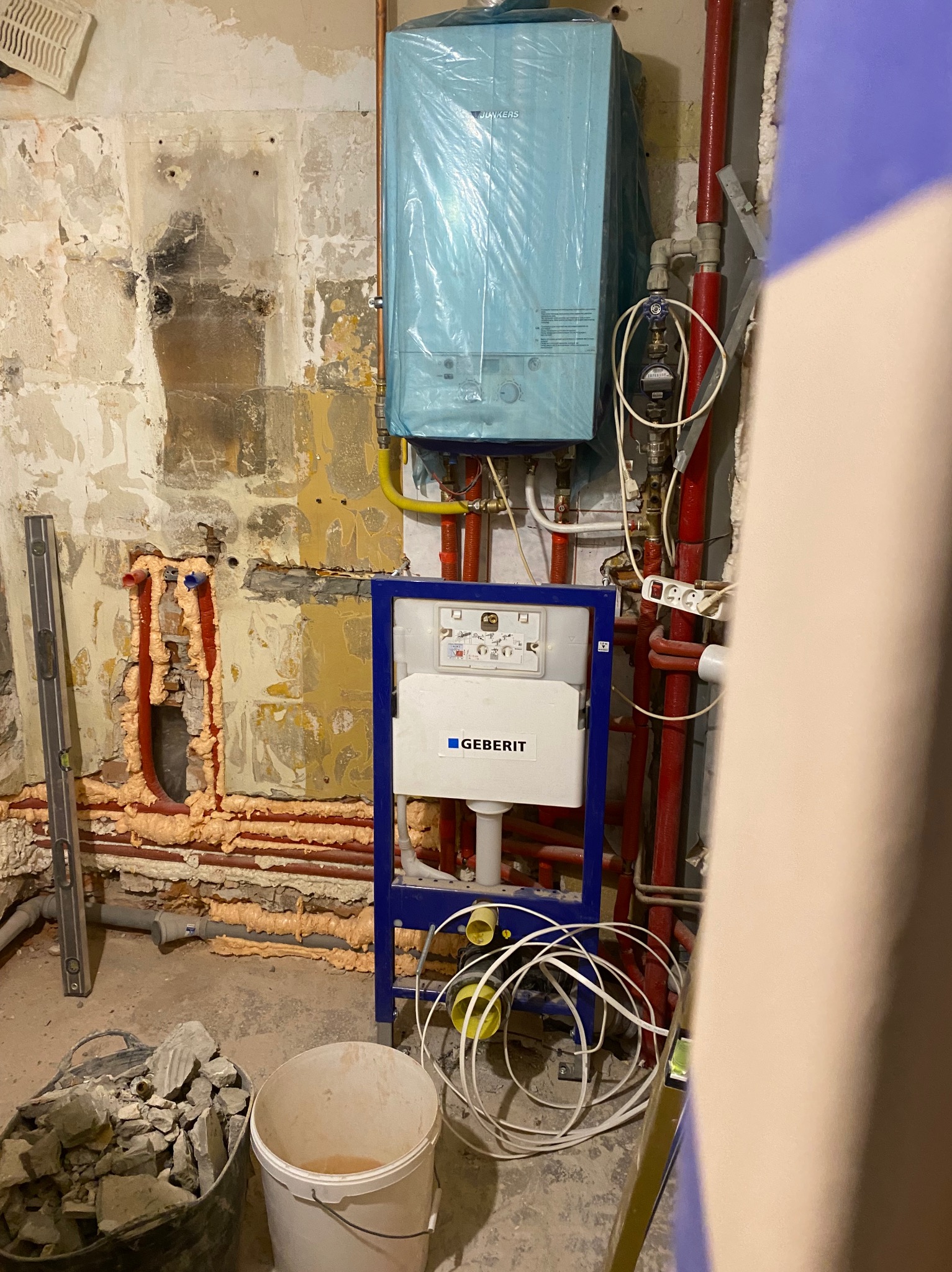 Instalacja podtynkowa Geberit w trakcie montażu, widoczne rury, pianka montażowa, poziomica i gruz, kocioł Junkers zabezpieczony folią.