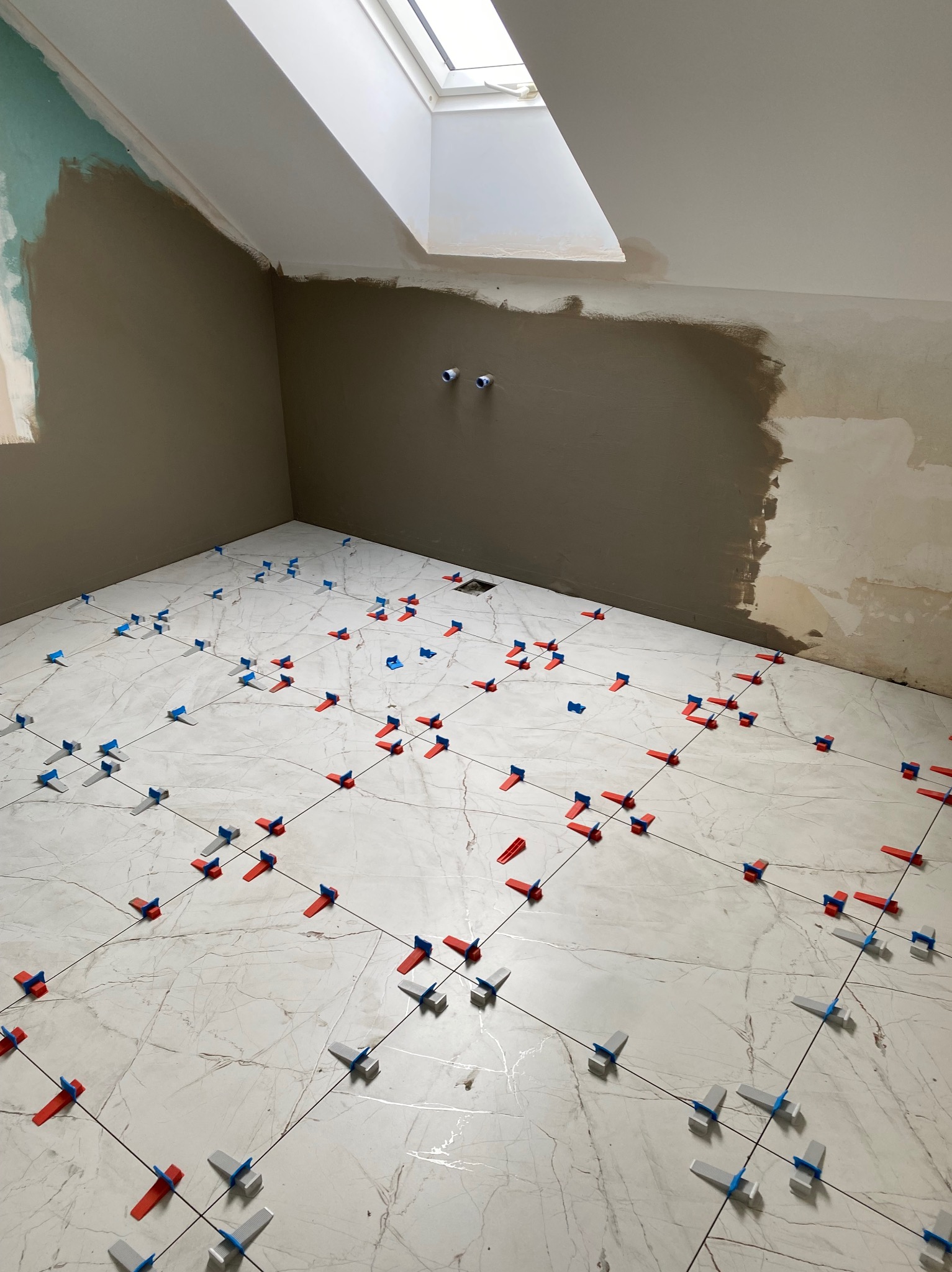 Podłoga w trakcie układania płytek imitujących marmur, z użyciem klipsów poziomujących w kolorach czerwonym, szarym i niebieskim, widoczne ślady zaprawy na ścianach i surowe wykończenie poddasza...