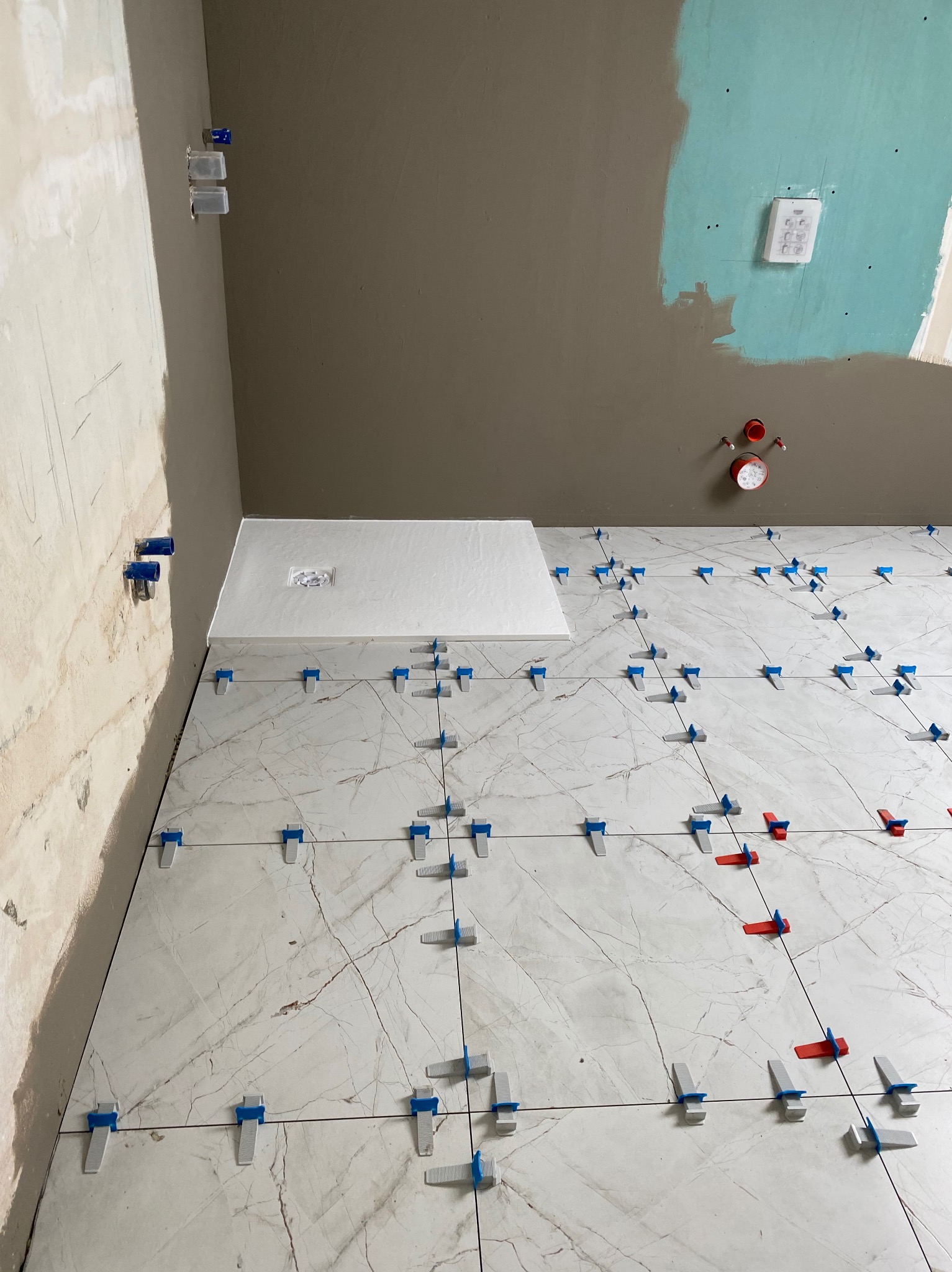 Układanie płytek podłogowych z marmurkowym wzorem, z użyciem niebieskich i czerwonych klipsów poziomujących, widoczne przygotowane ściany z instalacjami.