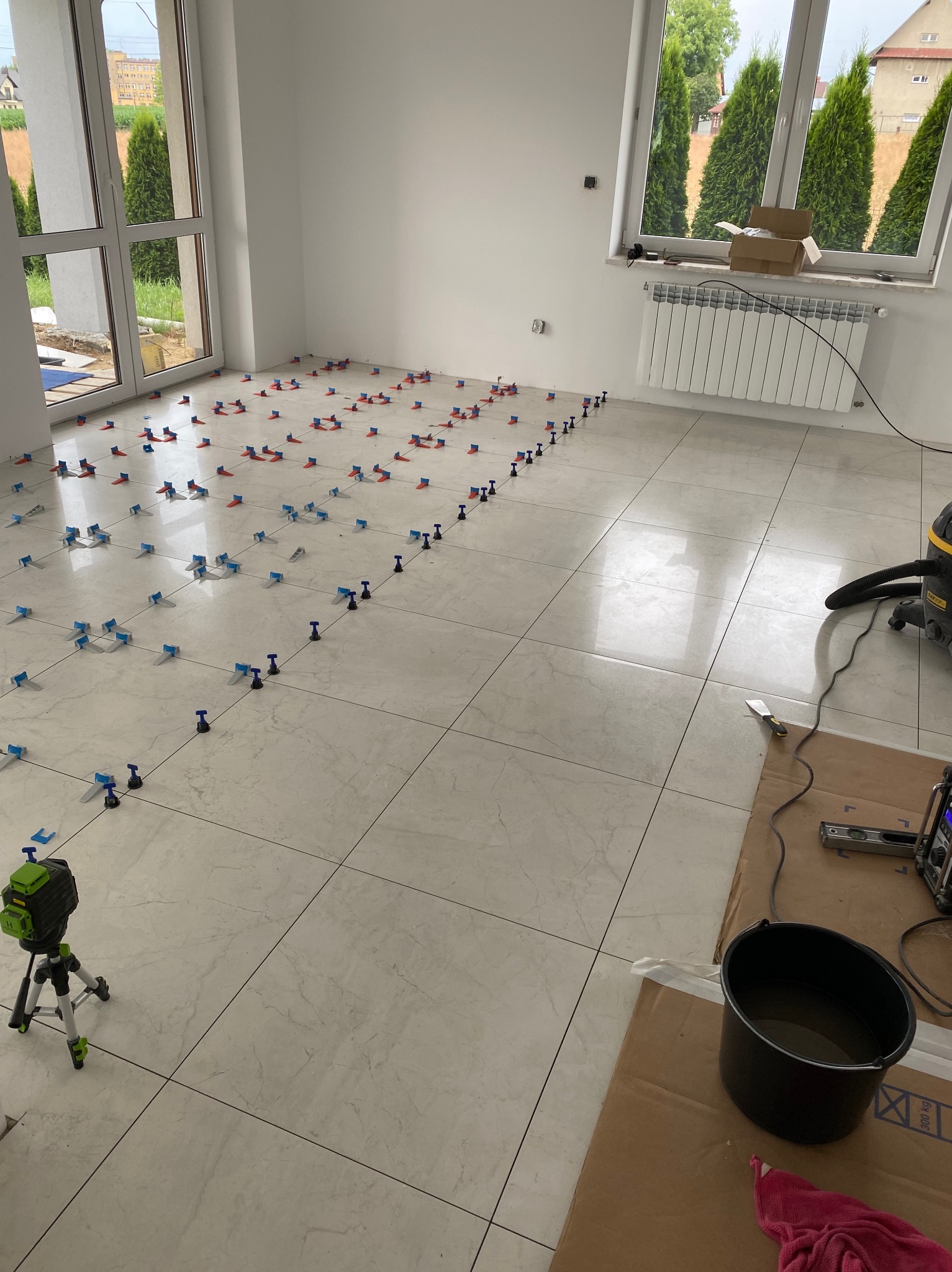 Jasne, prawie ukończone płytki podłogowe w pokoju z dużymi oknami, z systemem poziomowania płytek (klipsy i kliny) wzdłuż linii spoin, laser krzyżowy na statywie, wiadro z wodą i odkurzacz...