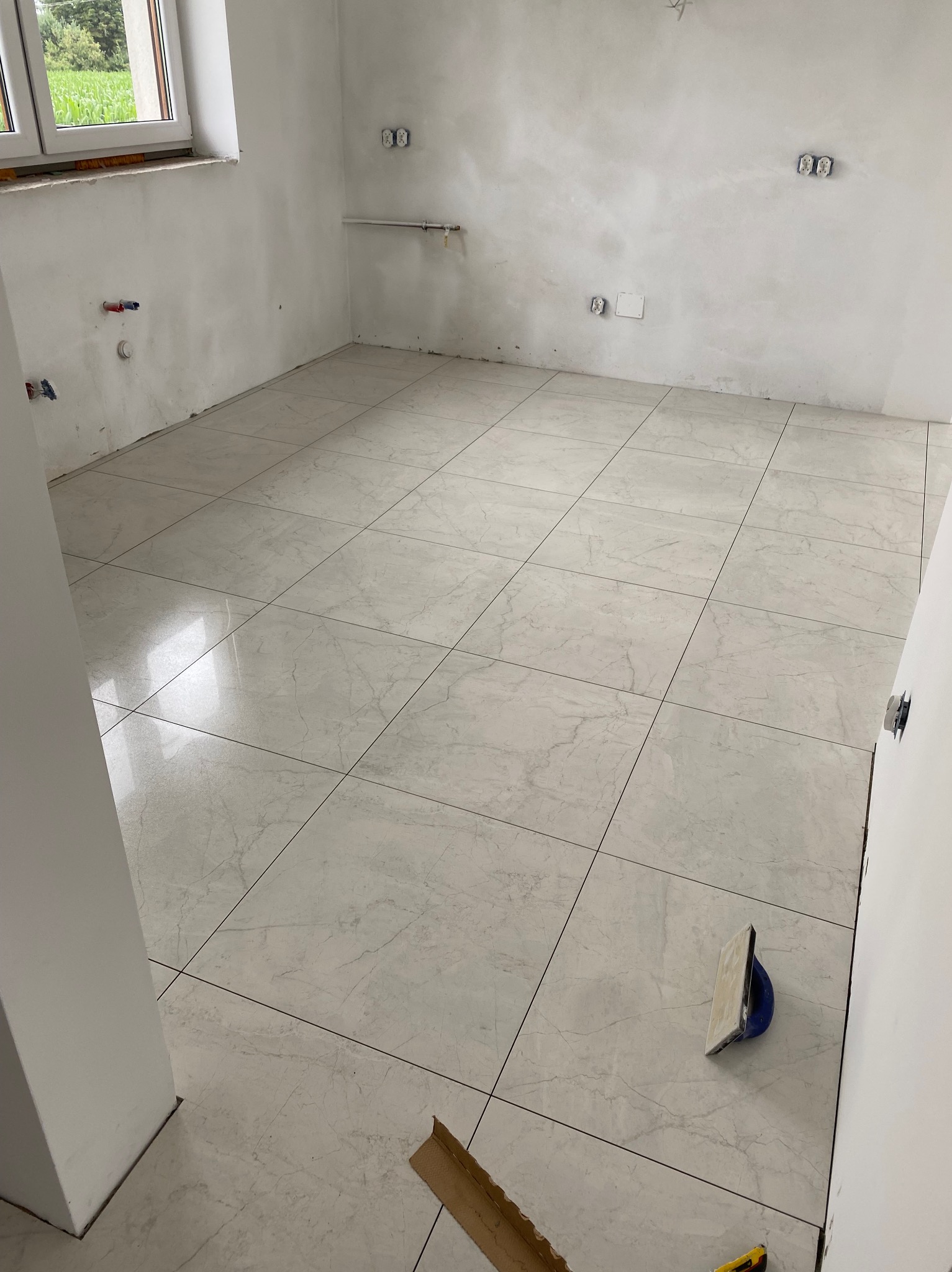 Ułożona podłoga z jasnych, marmurkowych płytek w pustym pomieszczeniu z widocznymi instalacjami elektrycznymi i narzędziami glazurniczymi.