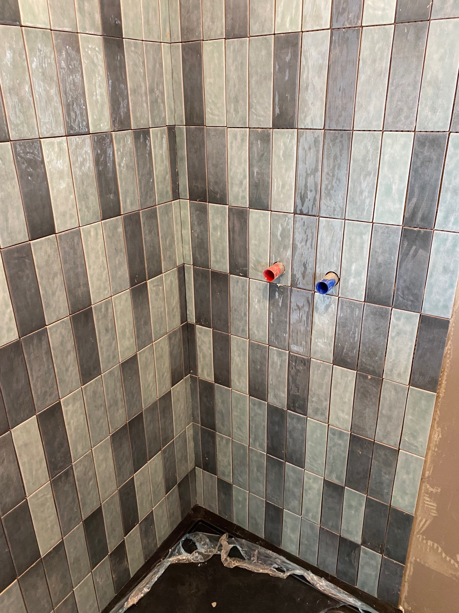 Ściana wyłożona pionowymi, prostokątnymi płytkami w odcieniach szarości i zieleni; widoczne wystające rury z instalacji wodnej w kolorach niebieskim i czerwonym, oraz folia ochronna na podłodze.