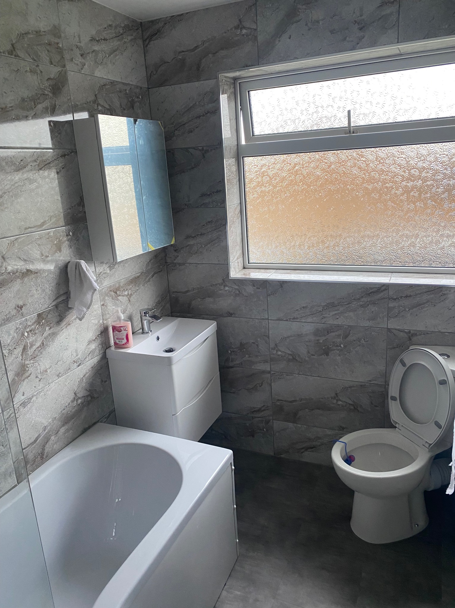 Wykończona łazienka z marmurowymi płytkami, białą armaturą, umywalką z szafką, wanną i toaletą z niebieskim odświeżaczem, oświetlona przez okno z mleczną szybą.