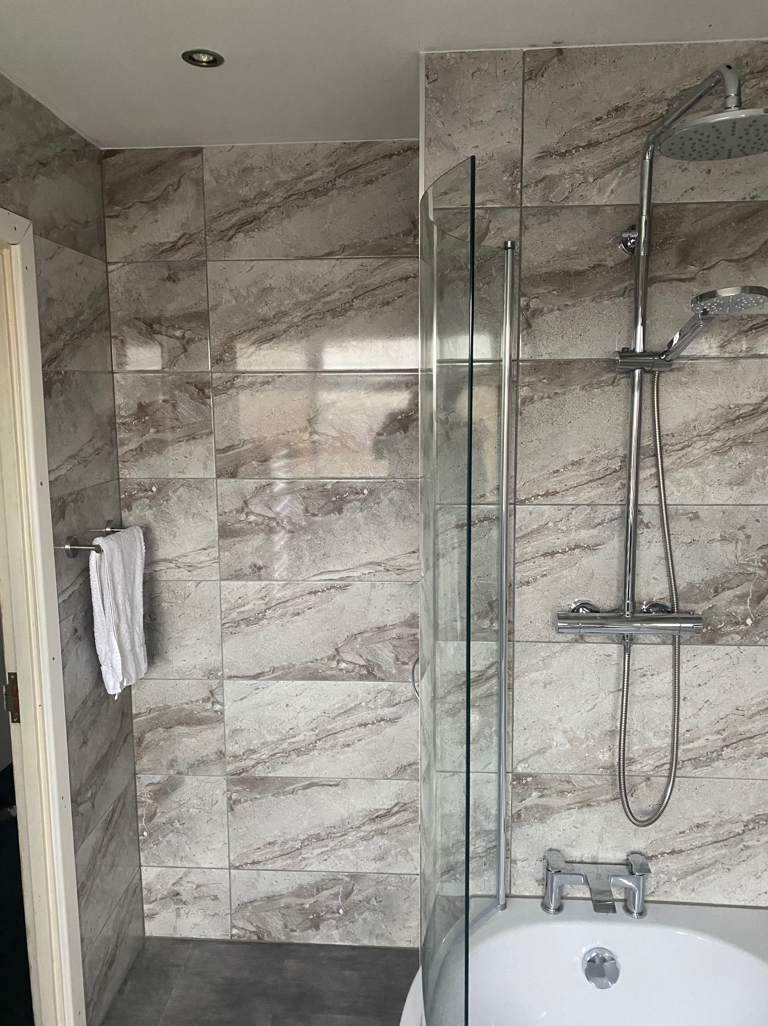 Wykończona łazienka z marmurowymi płytkami na ścianach, kabiną prysznicową ze szklanym panelem, chromowaną armaturą i białym ręcznikiem na wieszaku.
