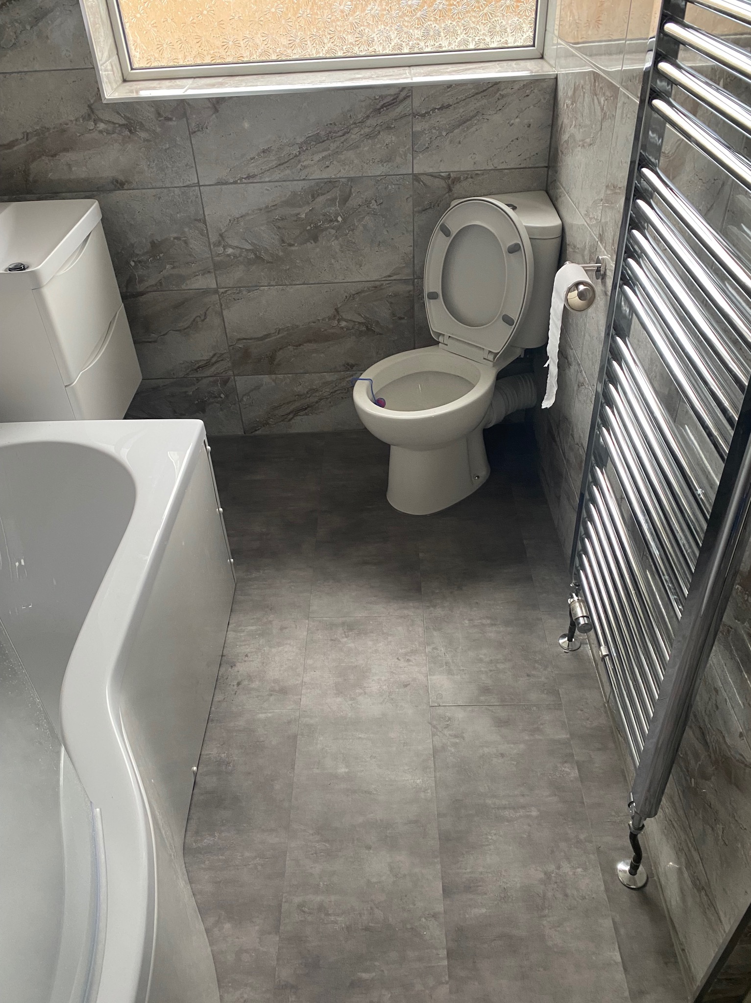 Ukończona łazienka z szarymi płytkami imitującymi marmur na ścianach i podłodze, biała wanna, toaleta z otwartą klapą, umywalka z szafką i chromowany grzejnik drabinkowy.
