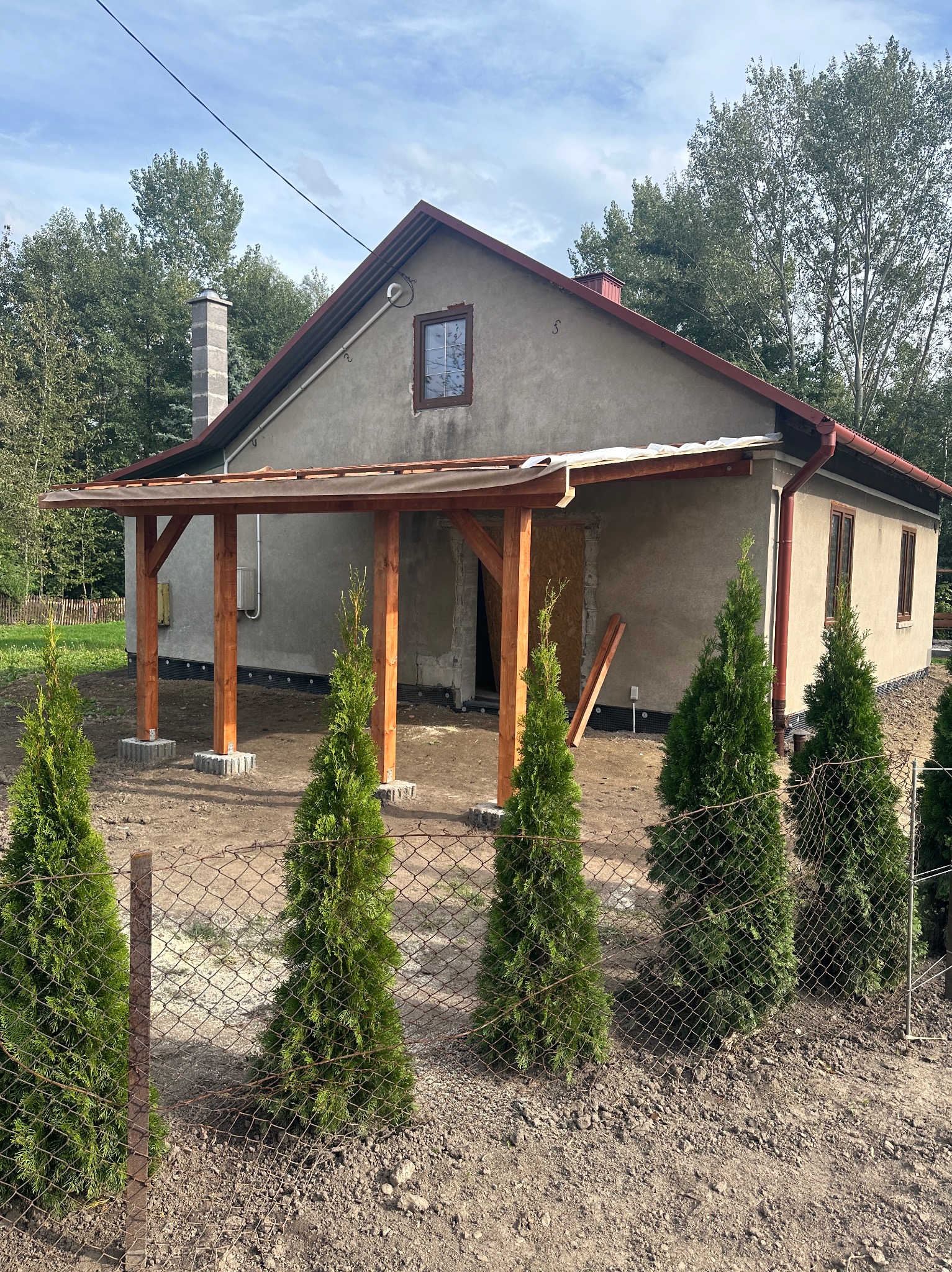 Drewniana konstrukcja zadaszenia nad wejściem do budynku w Grzęsce, widoczne słupy na betonowych podstawach i częściowo zamontowany dach, w tle surowa elewacja budynku i młode tuje wzdłuż ogrodzenia.