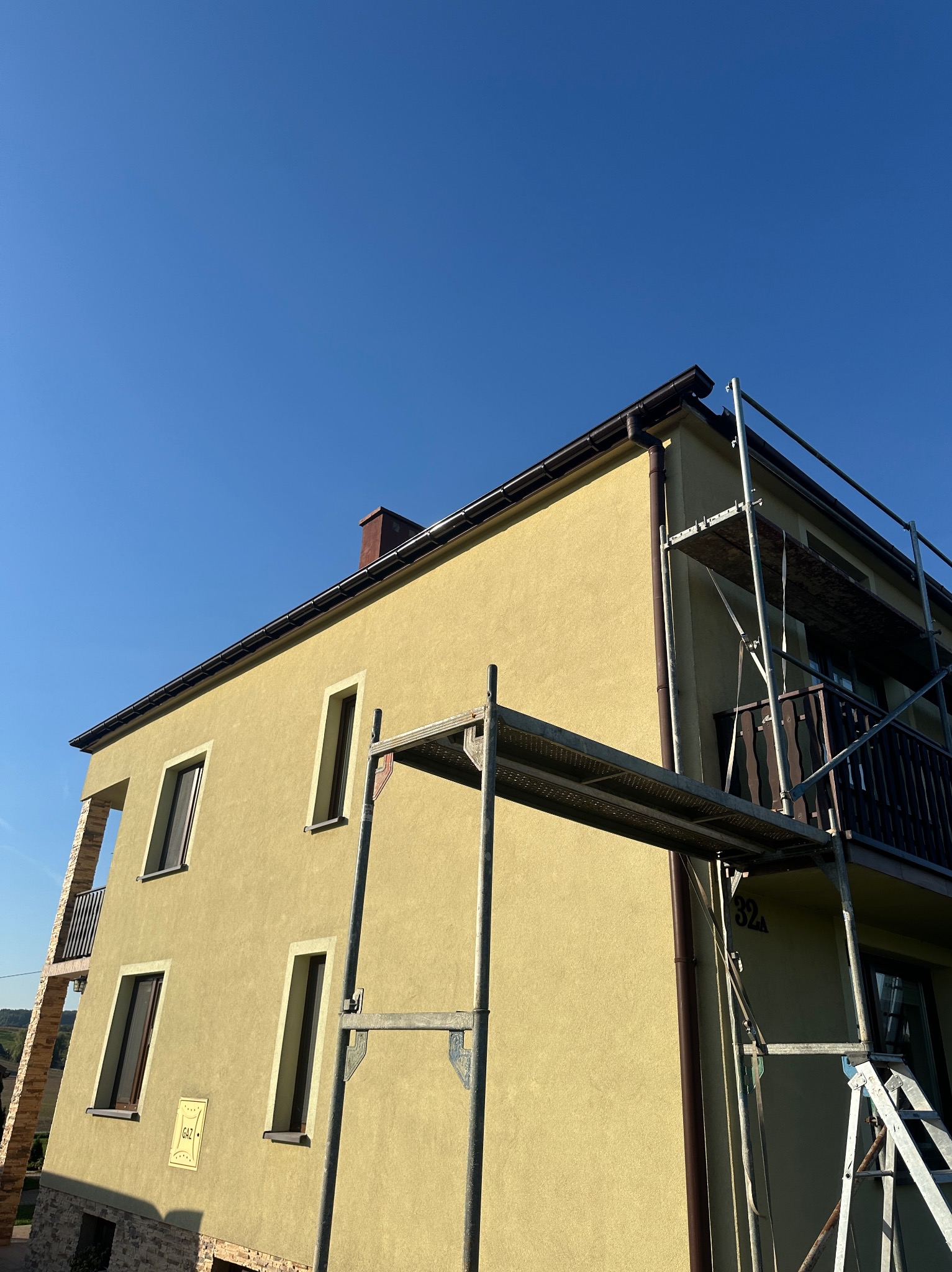 Żółty dom jednorodzinny z balkonem, w trakcie prac elewacyjnych, widoczne rusztowania i brązowe rynny, błękitne niebo w tle.