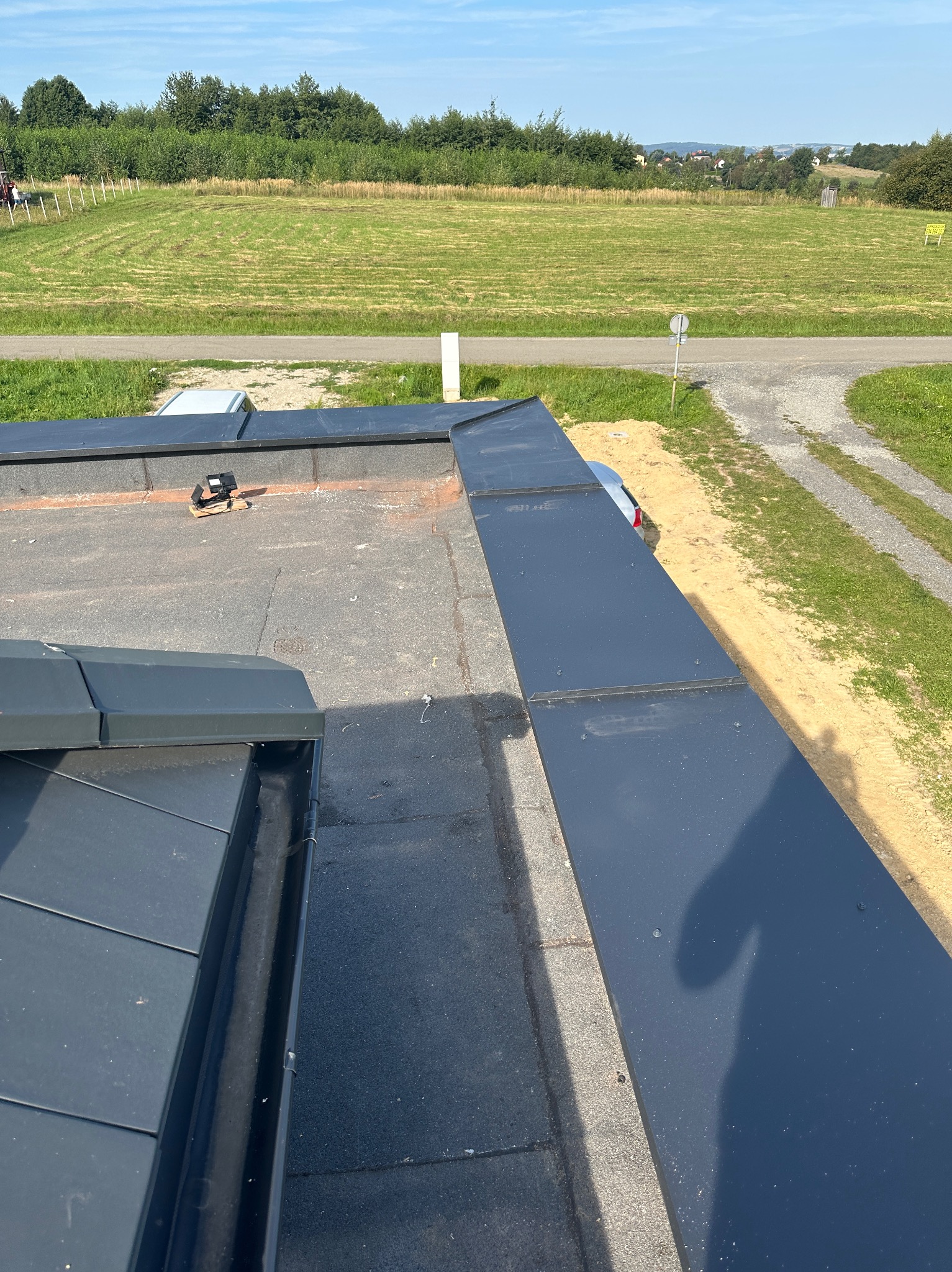 Nowe, grafitowe obróbki blacharskie na płaskim dachu, widoczne z góry, z narzędziami leżącymi obok, a w tle zielone pola i błękitne niebo.
