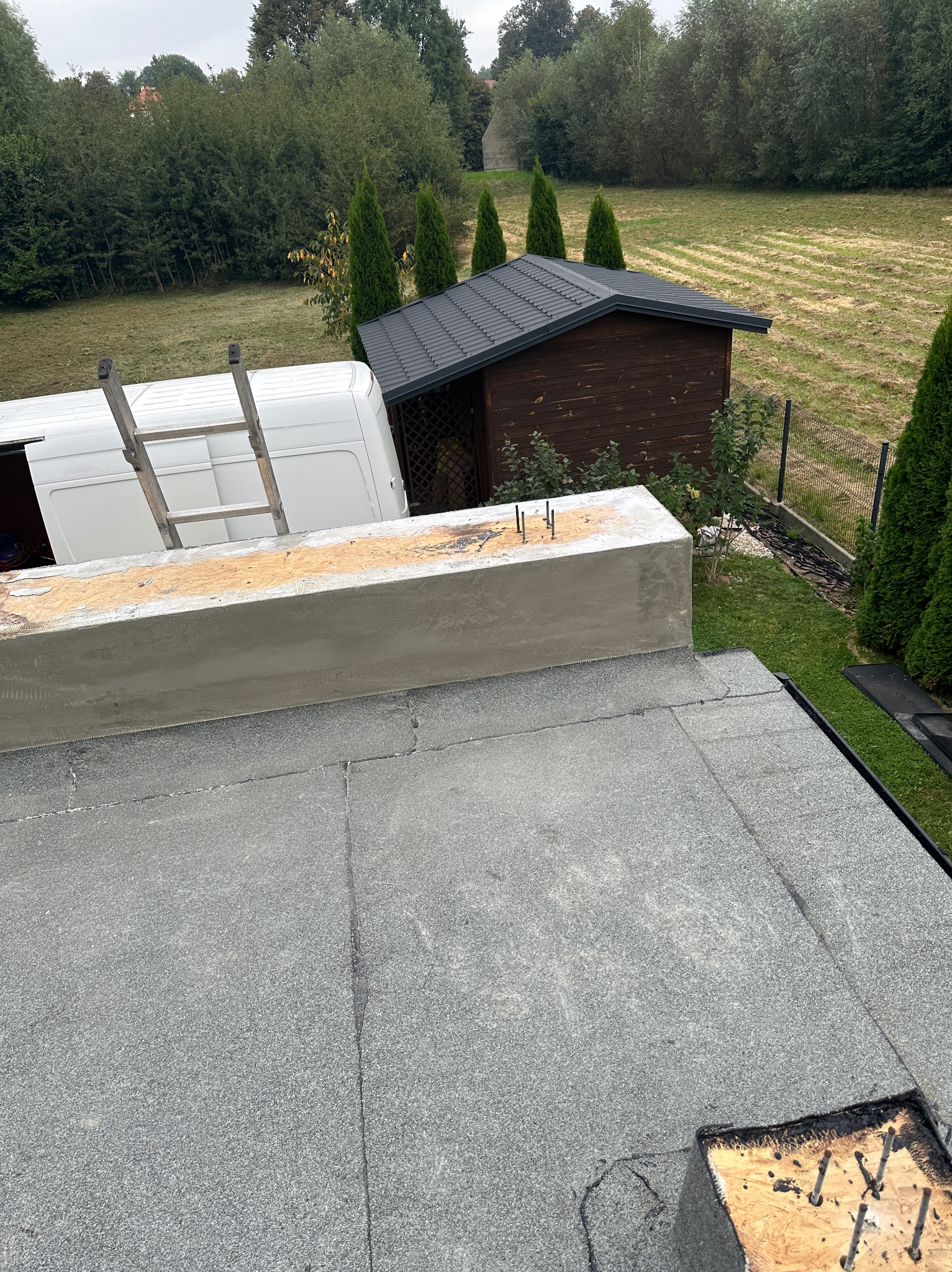 Widok z góry na płaski dach pokryty szarą papą, z widoczną obróbką blacharską przy krawędzi, fragmentem ściany attykowej z wystającymi prętami, białym samochodem dostawczym z drabiną w tle...