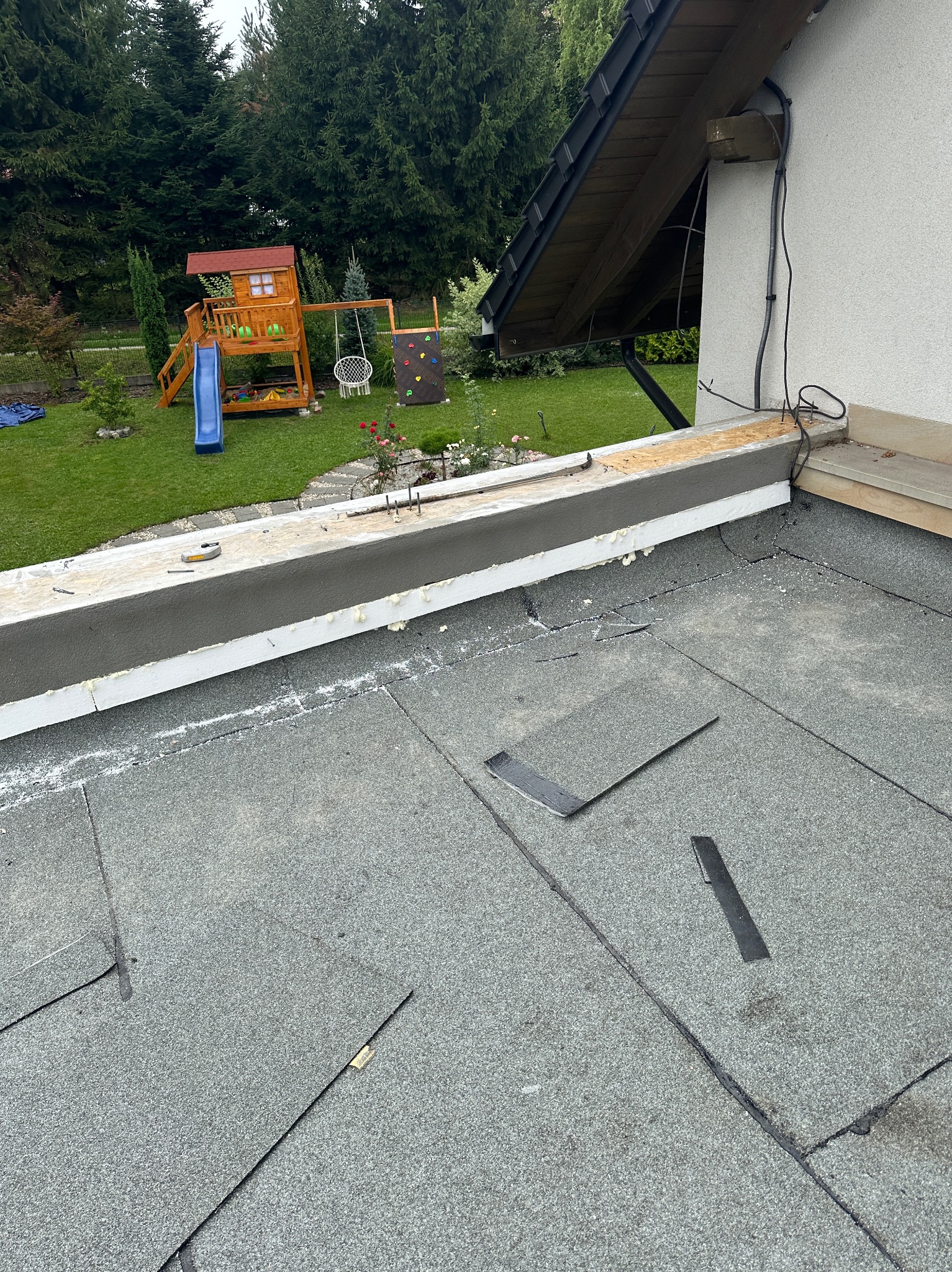 Fragment dachu pokrytego papą, z widocznymi łatami i narzędziami, w tle plac zabaw i dom z ogrodem.