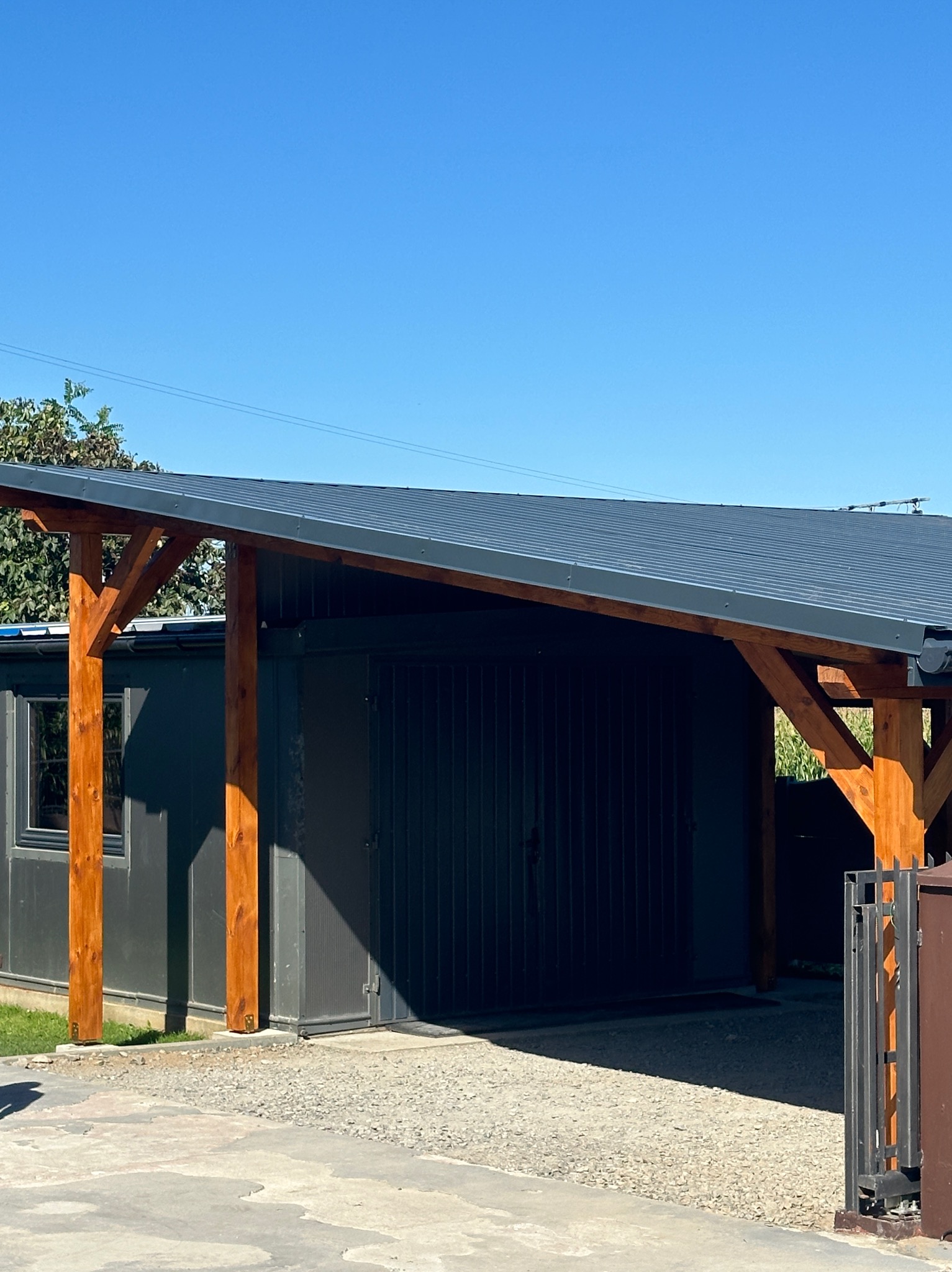 Zadaszenie nad stanowiskiem parkingowym, wsparte na drewnianych słupach, z blachy trapezowej w kolorze grafitowym, z widocznym fragmentem kontenera biurowego w tle.