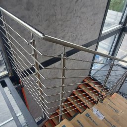 Alkord - Wnętrze budynku w trakcie wykańczania, widok z góry na schody z tymczasowymi okładzinami, zabezpieczone folią okna i balustradę ze stali nierdzewnej o minimalistycznym designie, na zewnątrz...