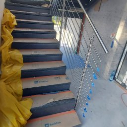 Alkord - Wewnętrzne schody z metalowymi stopniami zabezpieczonymi kartonem i folią, balustrada ze stali nierdzewnej z niebieskimi zaślepkami na końcach prętów.