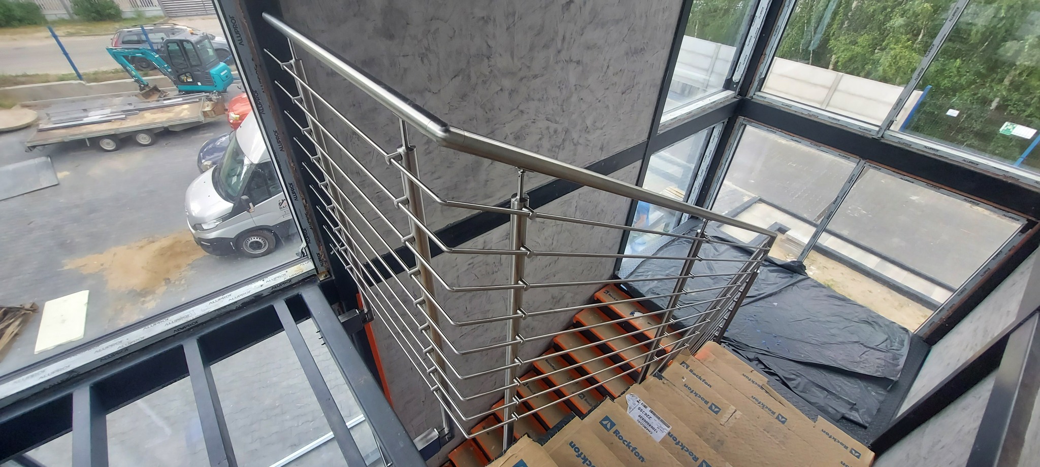 Wnętrze budynku w trakcie wykańczania, widok z góry na schody z tymczasowymi okładzinami, zabezpieczone folią okna i balustradę ze stali nierdzewnej o minimalistycznym designie, na zewnątrz...