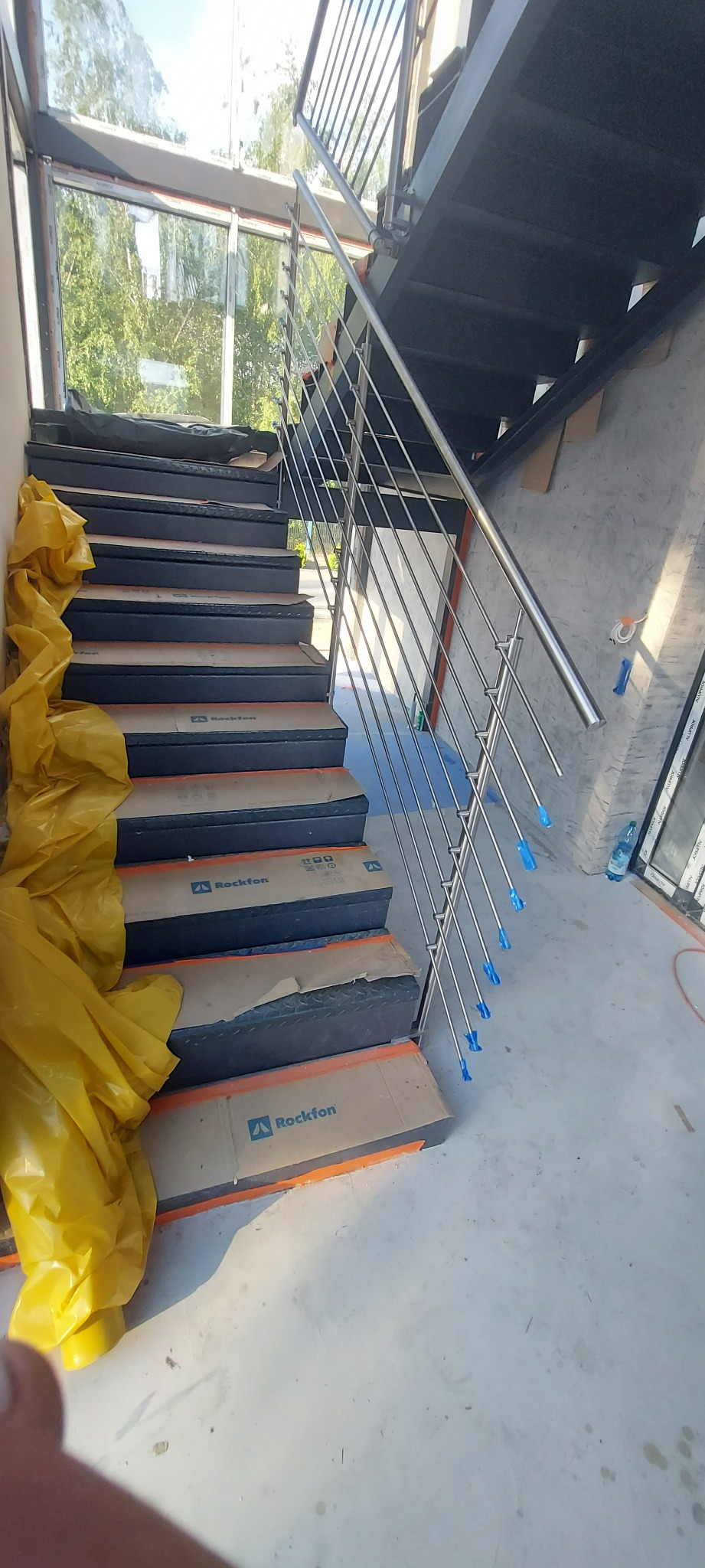 Wewnętrzne schody z metalowymi stopniami zabezpieczonymi kartonem i folią, balustrada ze stali nierdzewnej z niebieskimi zaślepkami na końcach prętów.