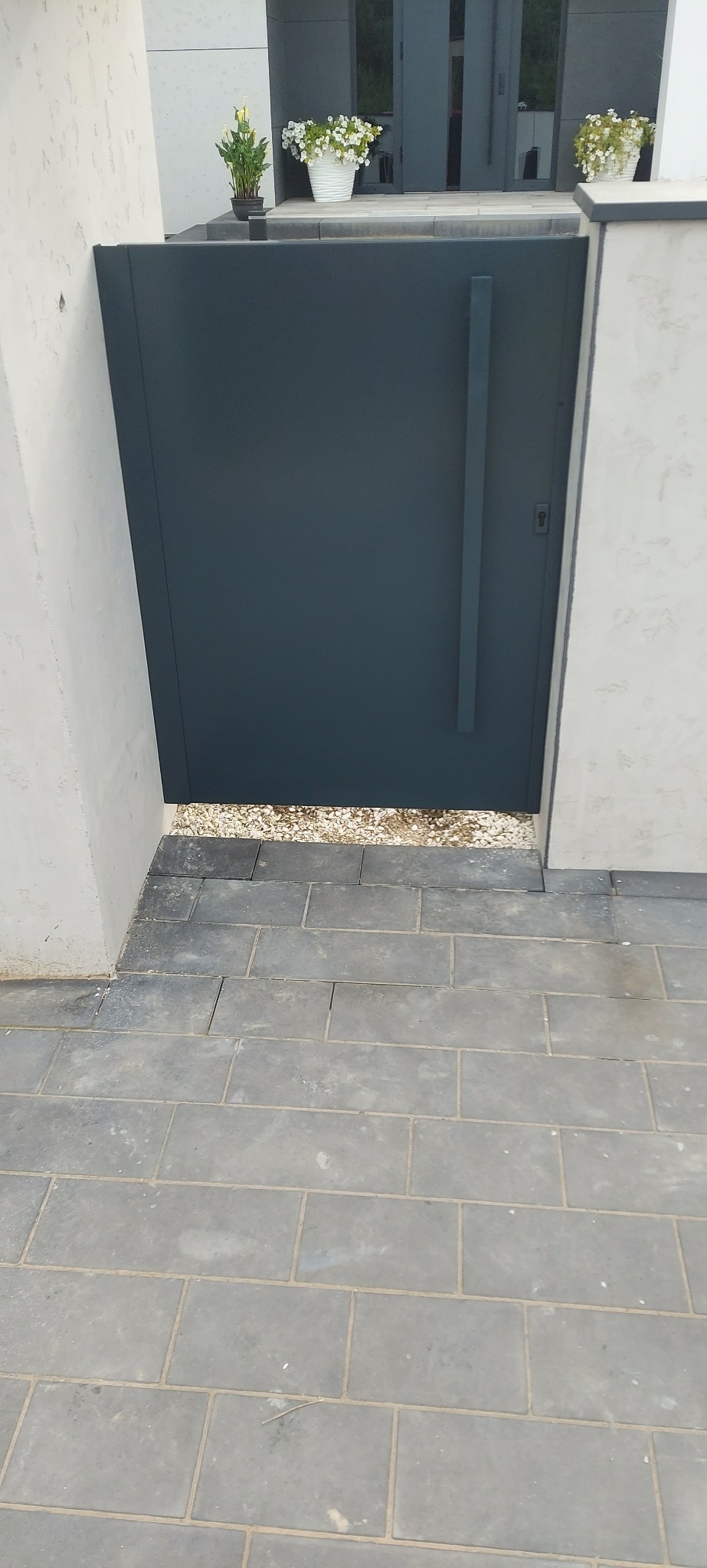 Nowoczesna, minimalistyczna brama wjazdowa w kolorze grafitowym, z pionowym uchwytem, wkomponowana w jasny mur, z widocznymi kwiatami w donicach na górze i szarą kostką brukową na dole.