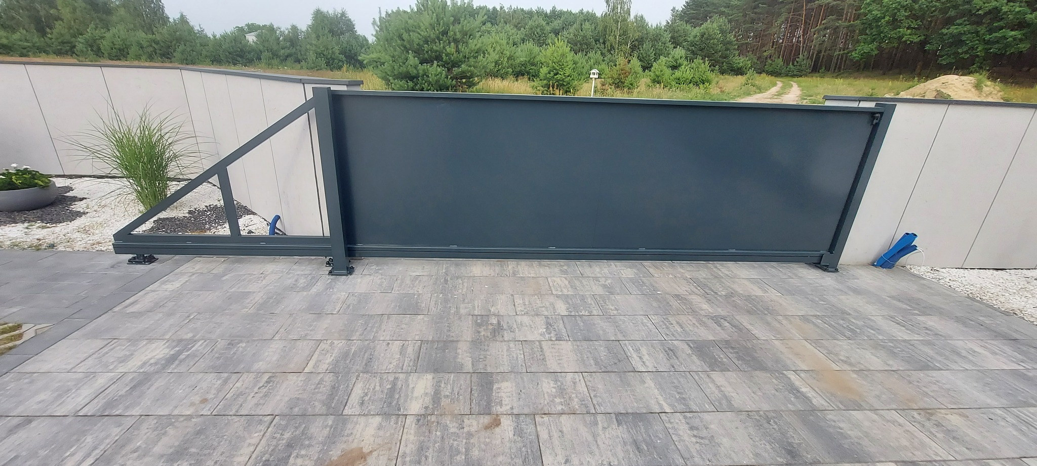 Nowoczesna, przesuwna brama wjazdowa w kolorze grafitowym, z widocznym mechanizmem jezdnym, zamontowana pomiędzy dwoma białymi, ukośnymi panelami ogrodzeniowymi, na tle zieleni drzew.