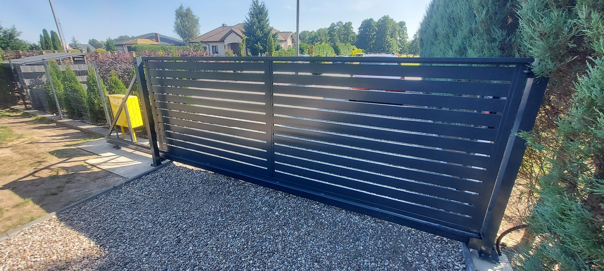 Nowoczesna, przesuwna brama wjazdowa z poziomymi panelami w kolorze antracytowym, zamontowana na posesji z podjazdem wysypanym żwirem, z widocznym fragmentem ogrodzenia i zieleni w tle.