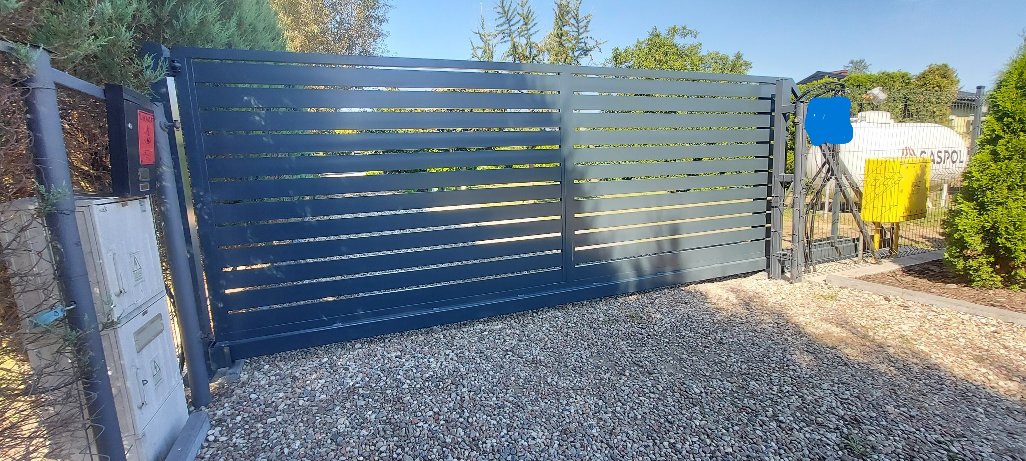 Nowoczesna przesuwna brama wjazdowa z poziomymi panelami w kolorze grafitowym, widoczna z bliska na żwirowym podłożu, z instalacją gazową w tle.
