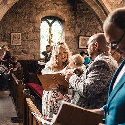ZoomArt - Wnętrze kościoła z kamiennymi ścianami, gdzie goście ślubni czytają z modlitewników, w centrum matka z dzieckiem spogląda na otwartą księgę.