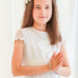 ZoomArt - Dziewczynka w białej sukience komunijnej z wiankiem na głowie i różańcem w dłoniach, złożonych do modlitwy na tle białej zasłony.