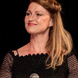 ZoomArt - Uśmiechnięta kobieta z brokatowymi piegami, trzymająca mikrofon, w czarnej, błyszczącej sukience z koronkowymi rękawami, na czarnym tle.