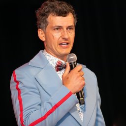 ZoomArt - Mężczyzna w błękitnym garniturze z czerwoną lamówką, muszką w kratę i brokatem na twarzy trzyma mikrofon na czarnym tle.