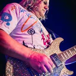 ZoomArt - Muzyk z długimi blond włosami gra na błyszczącej, złotej gitarze elektrycznej w trakcie koncertu, ujęcie od dołu w mocnym oświetleniu scenicznym.