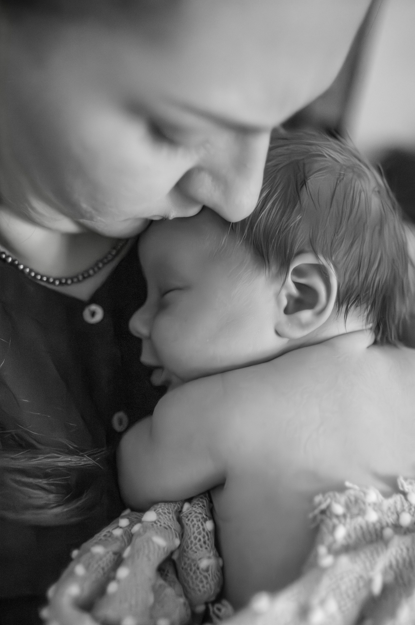 Czarno-białe zbliżenie matki całującej śpiące niemowlę w czoło, dziecko owinięte w dziergany kocyk w kropki.