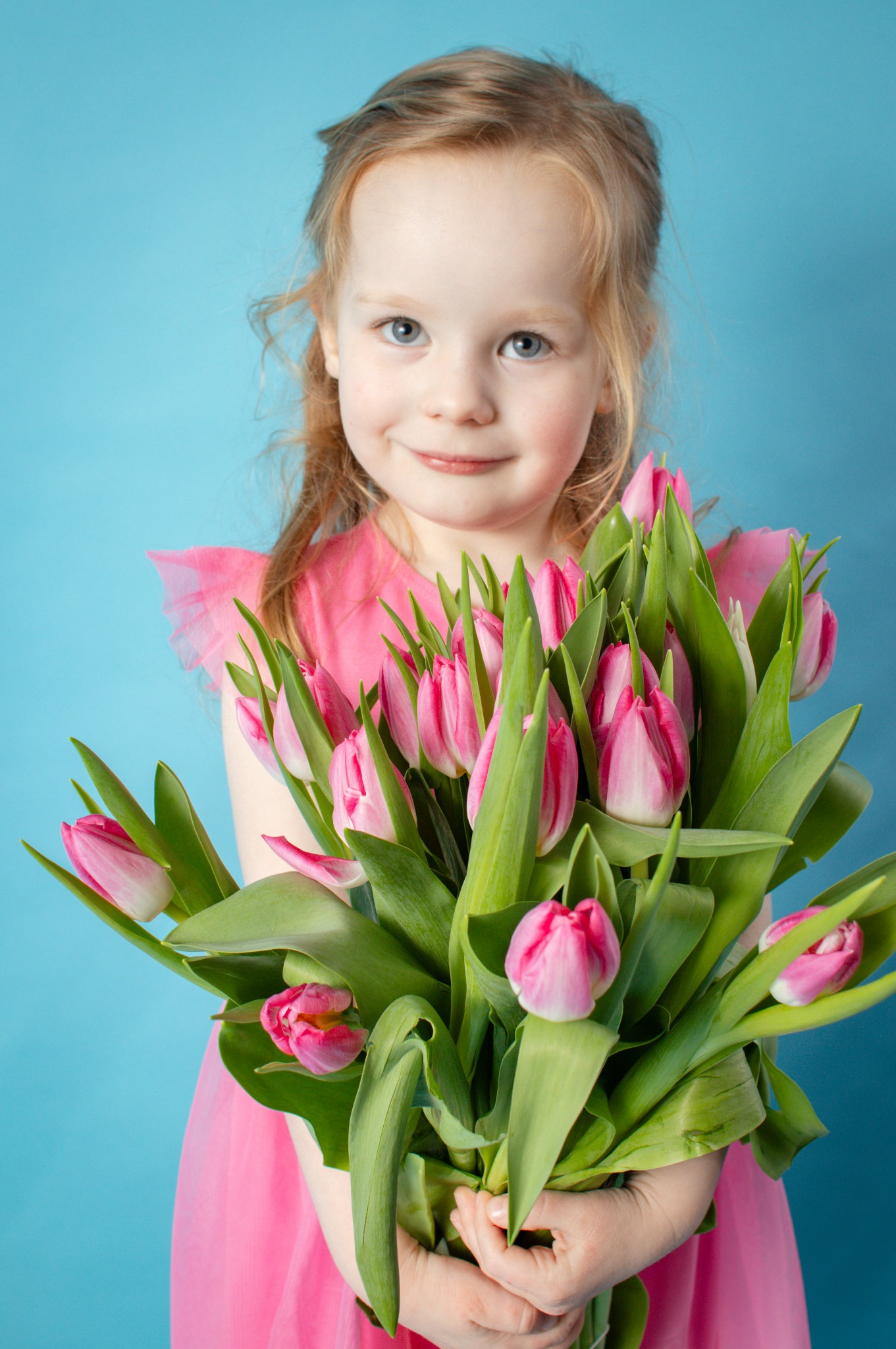 Uśmiechnięta dziewczynka w różowej sukience trzyma bukiet różowych tulipanów na tle błękitnego studia.