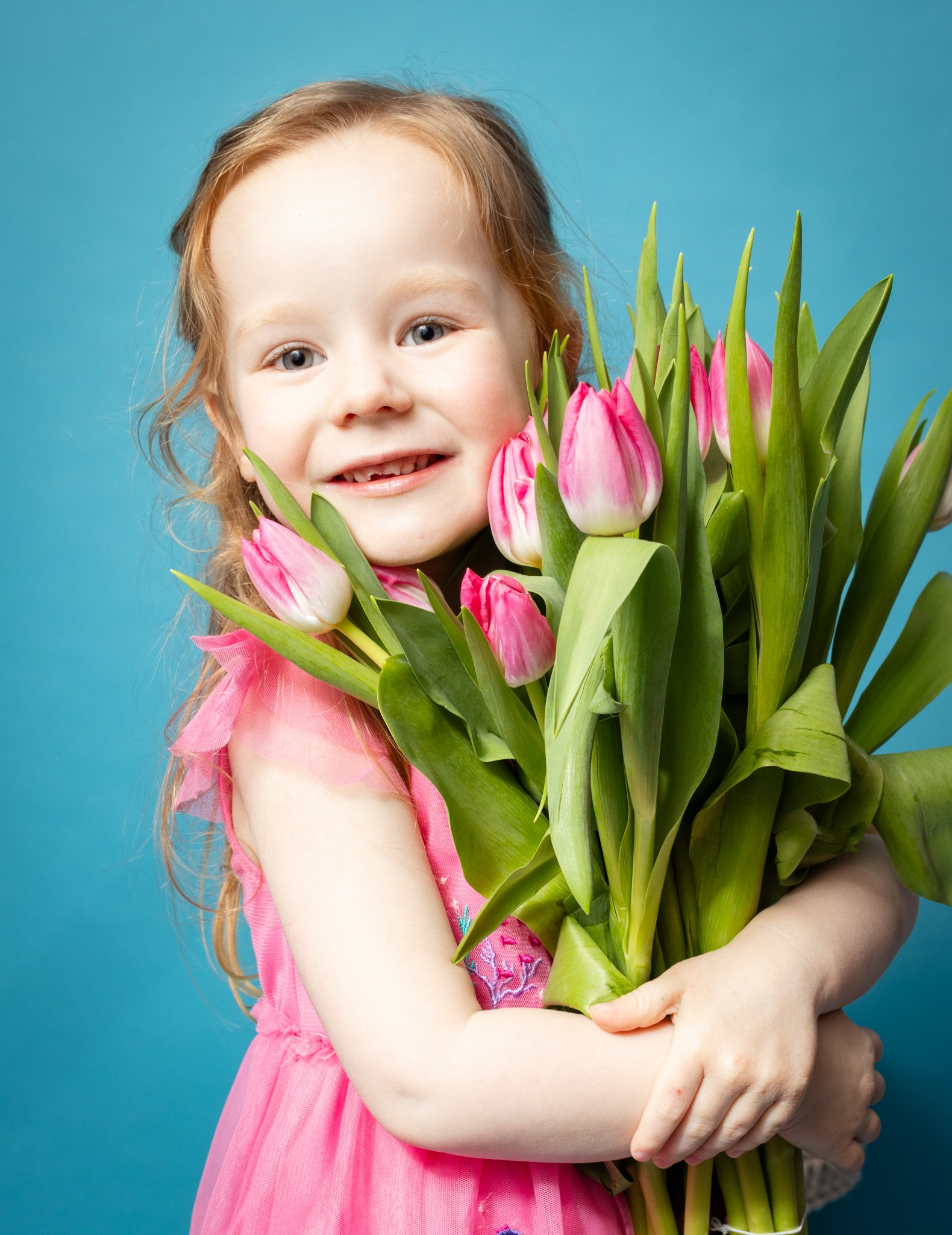 Uśmiechnięta dziewczynka w różowej sukience trzyma bukiet różowych tulipanów na turkusowym tle.